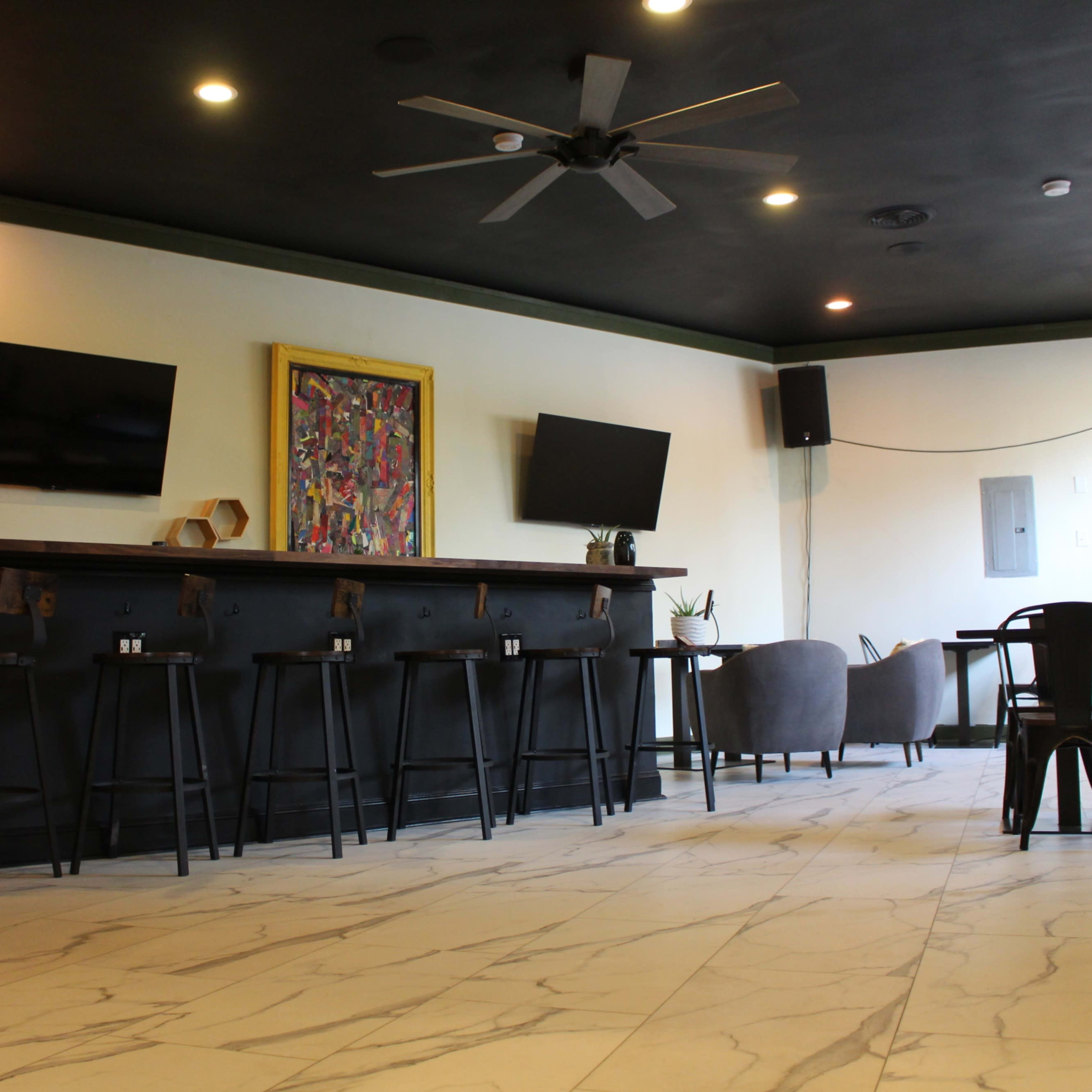 A modern bar area with a long black countertop, several bar stools, two wall-mounted televisions, and a seating area in a room with tiled flooring and dark ceiling.