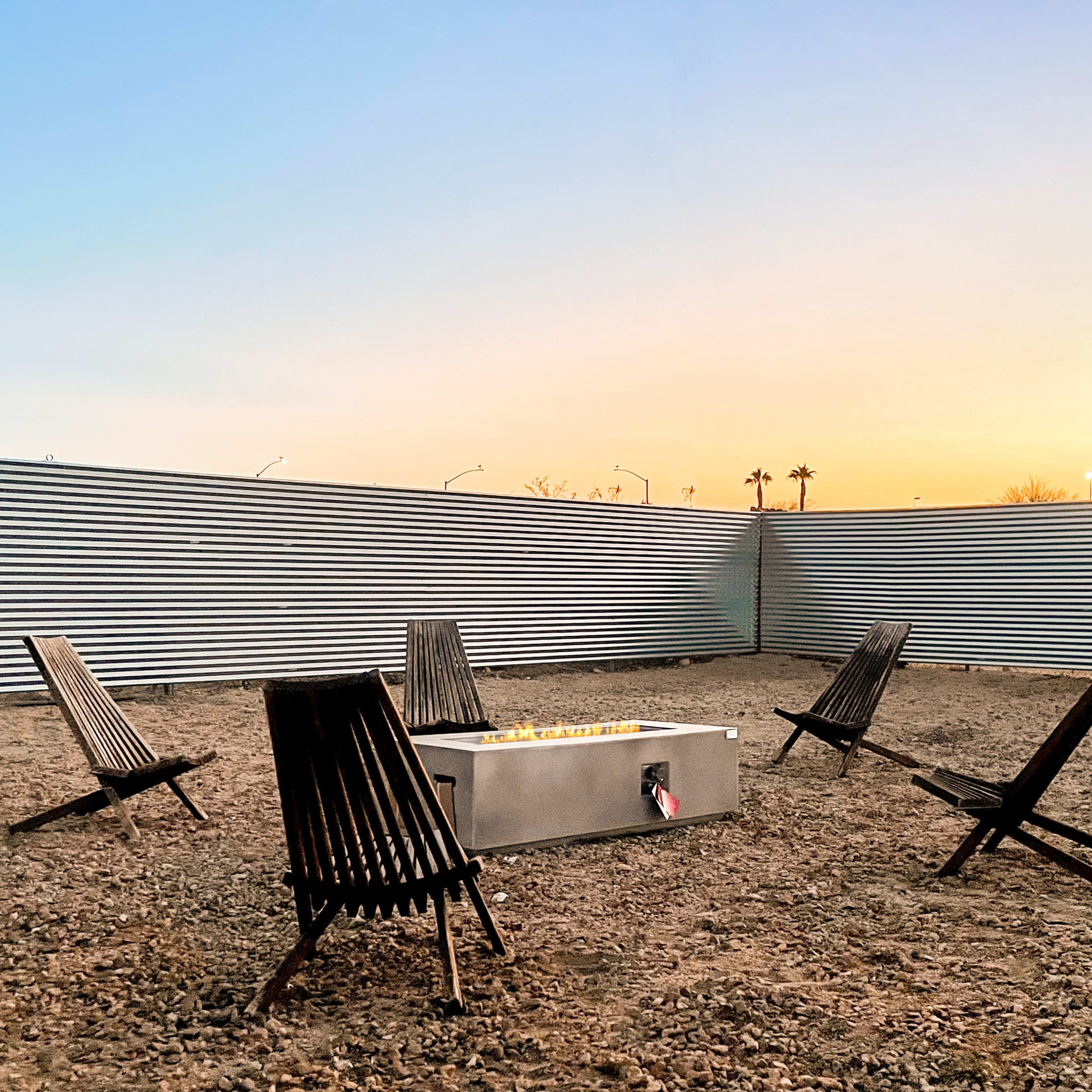 A circular arrangement of wooden chairs surrounds a metal fire pit in a fenced outdoor space at sunset.