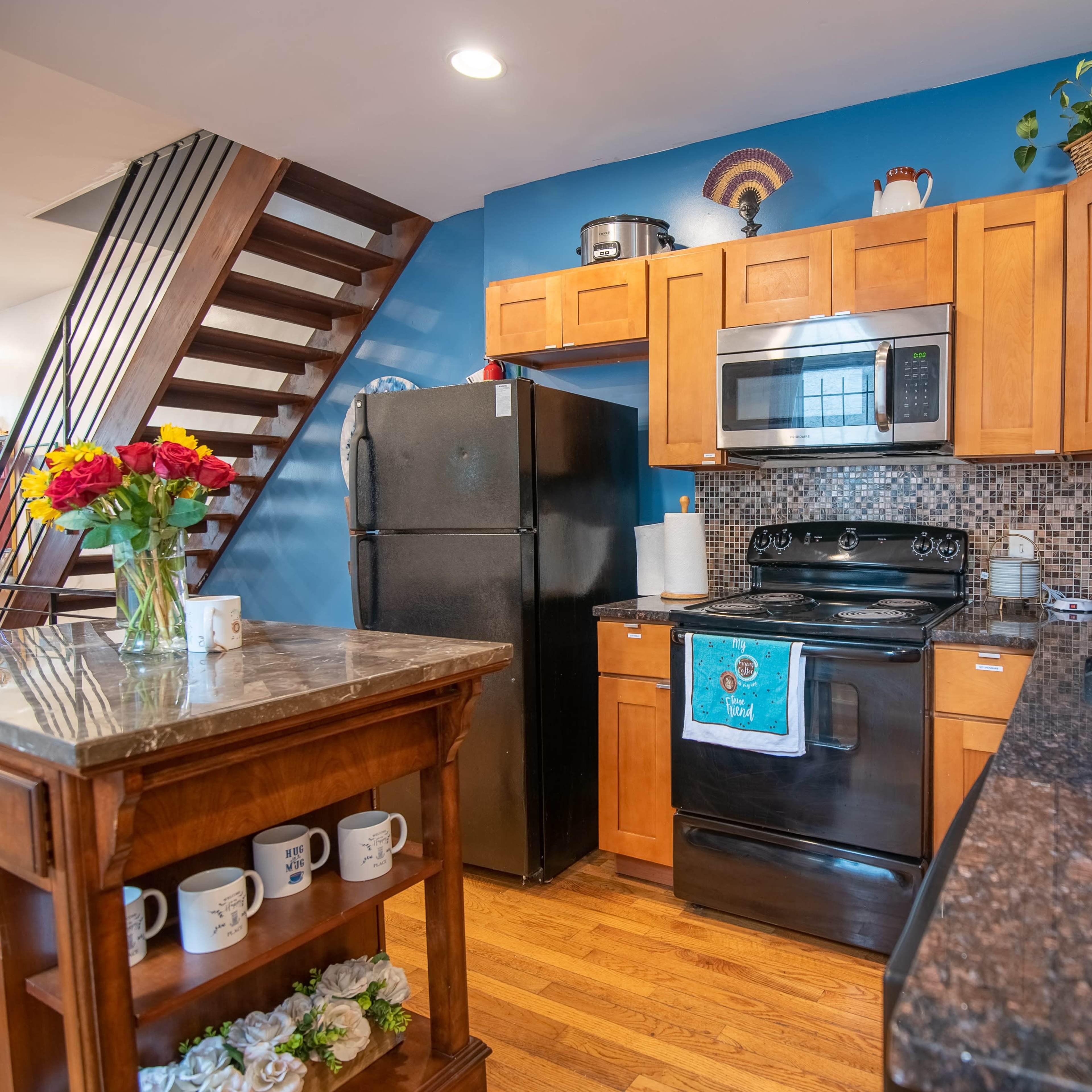 A modern kitchen features wooden cabinets, a black refrigerator, and a stove, with a granite countertop and a vase of flowers on the table.
