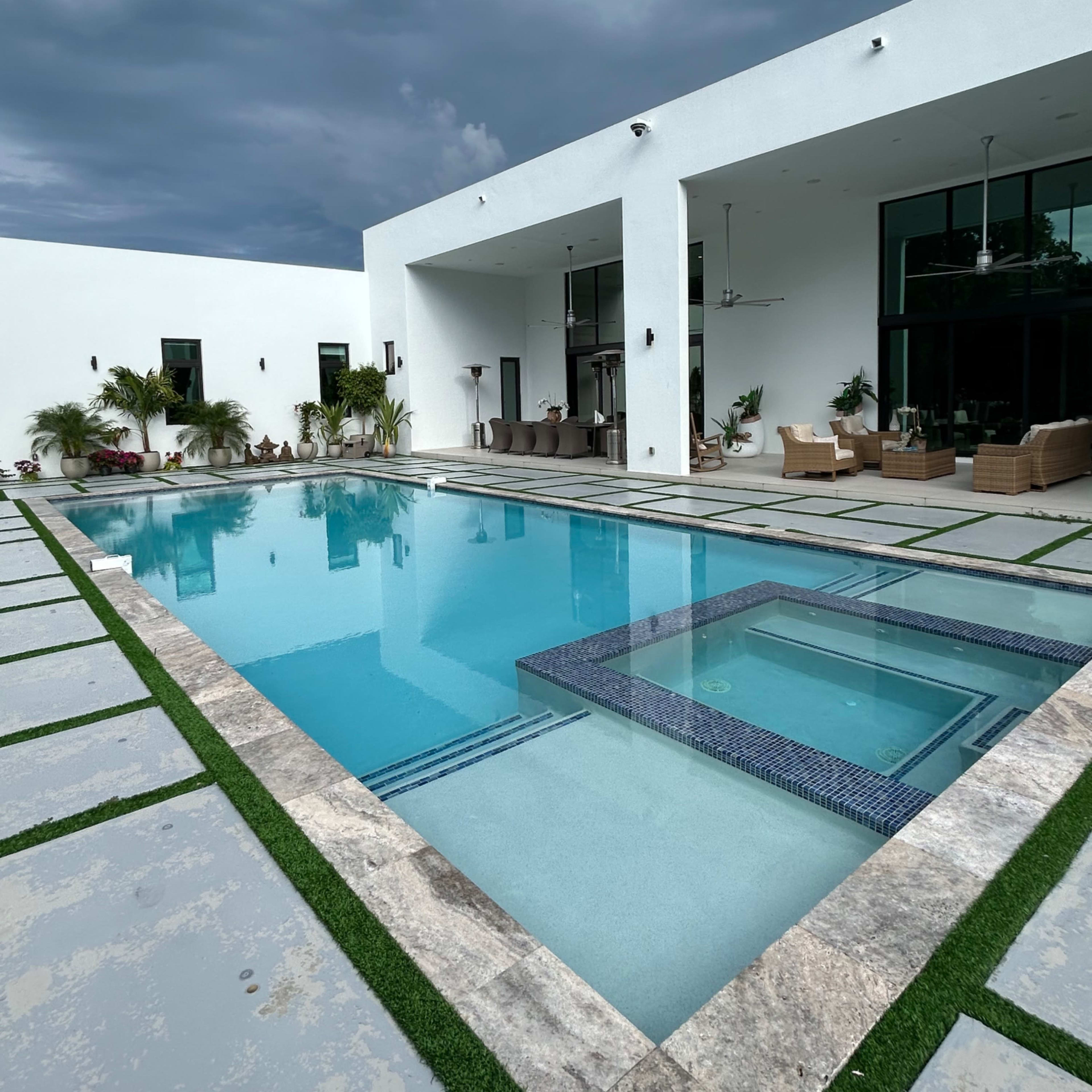 A modern house features a rectangular swimming pool surrounded by stone tiles and grass, with large windows and a covered seating area in the background.