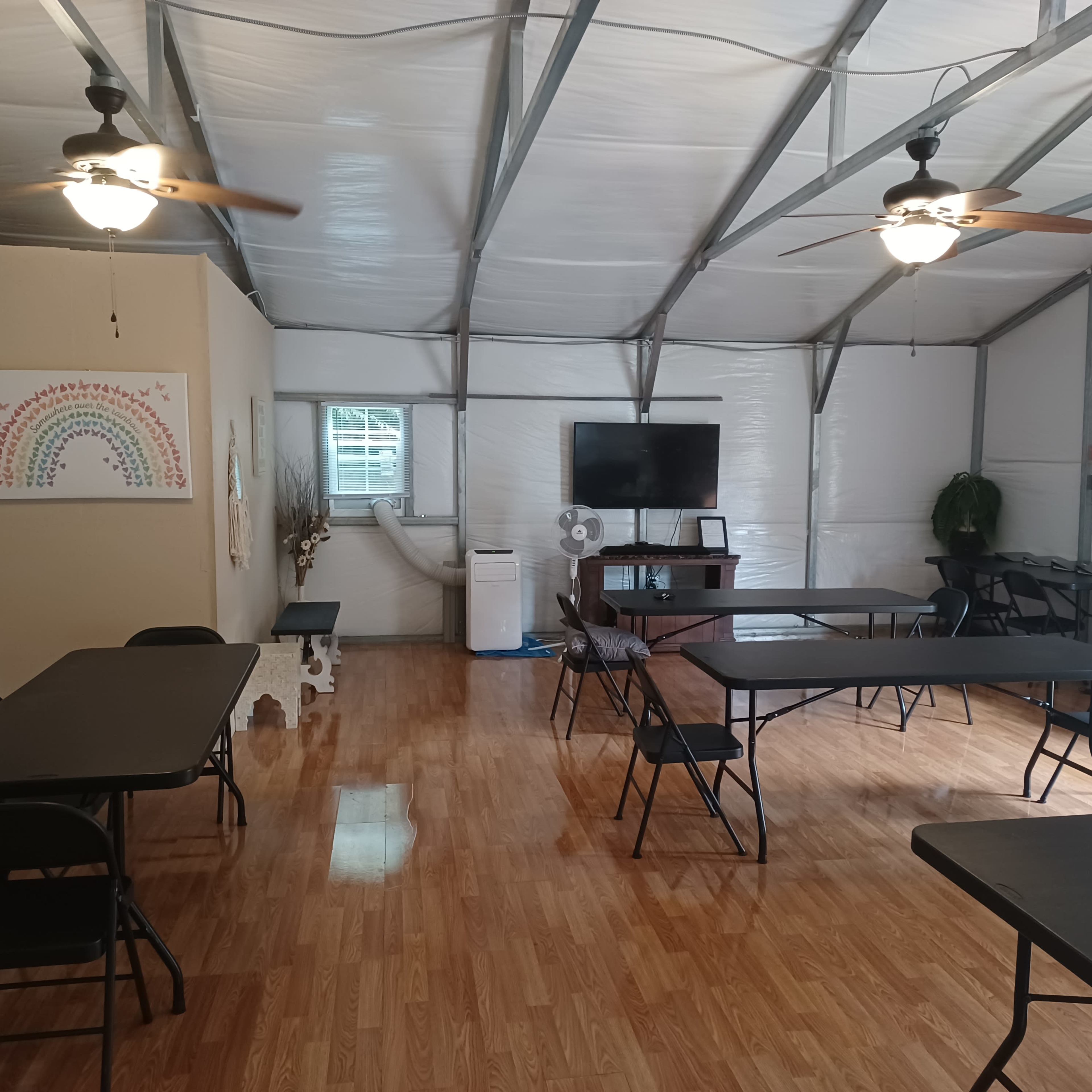 The interior of a spacious room features several black folding tables and chairs, a large television mounted on the wall, and a decorative rainbow painting.