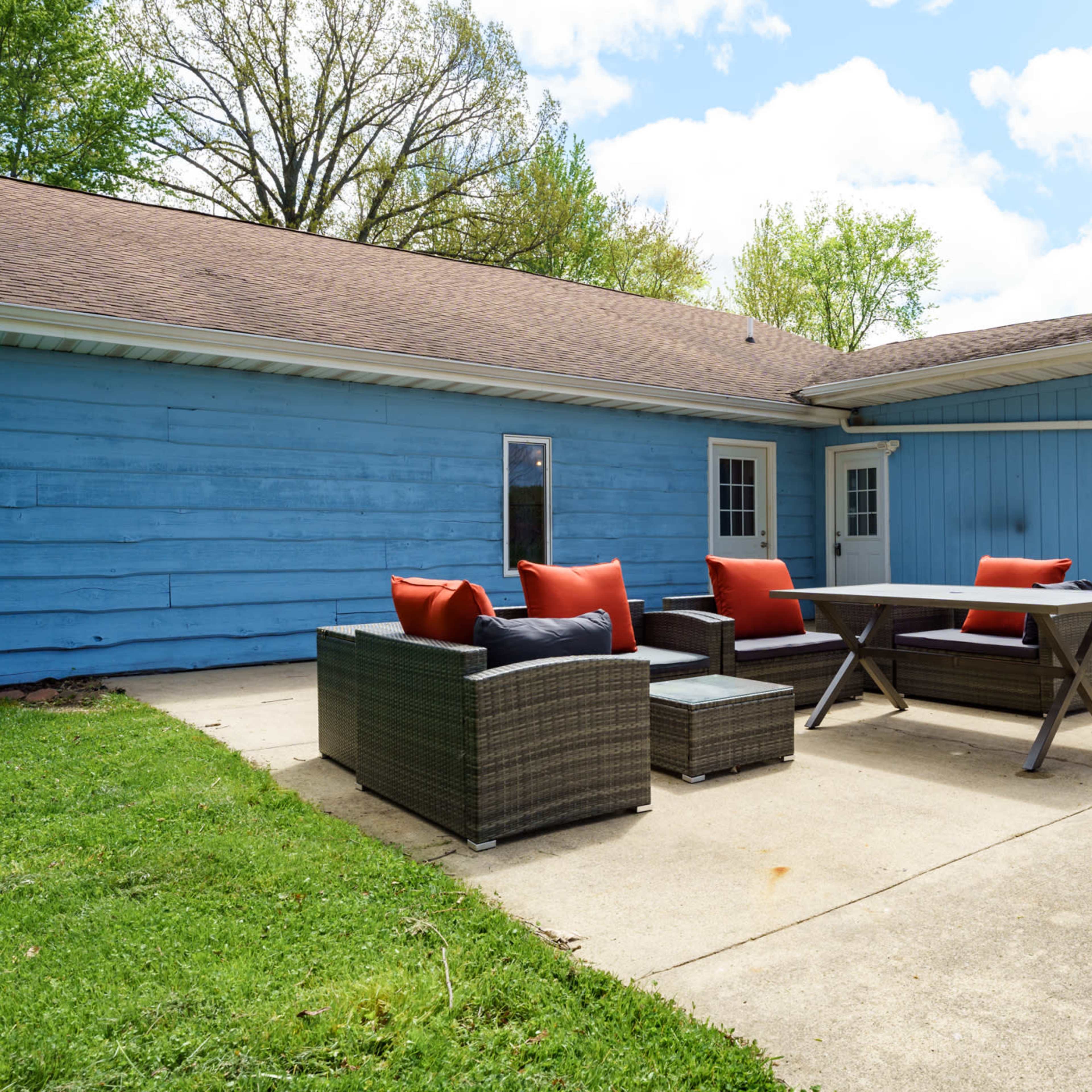 A patio area features a table and chairs surrounded by green grass and a blue house in the background.