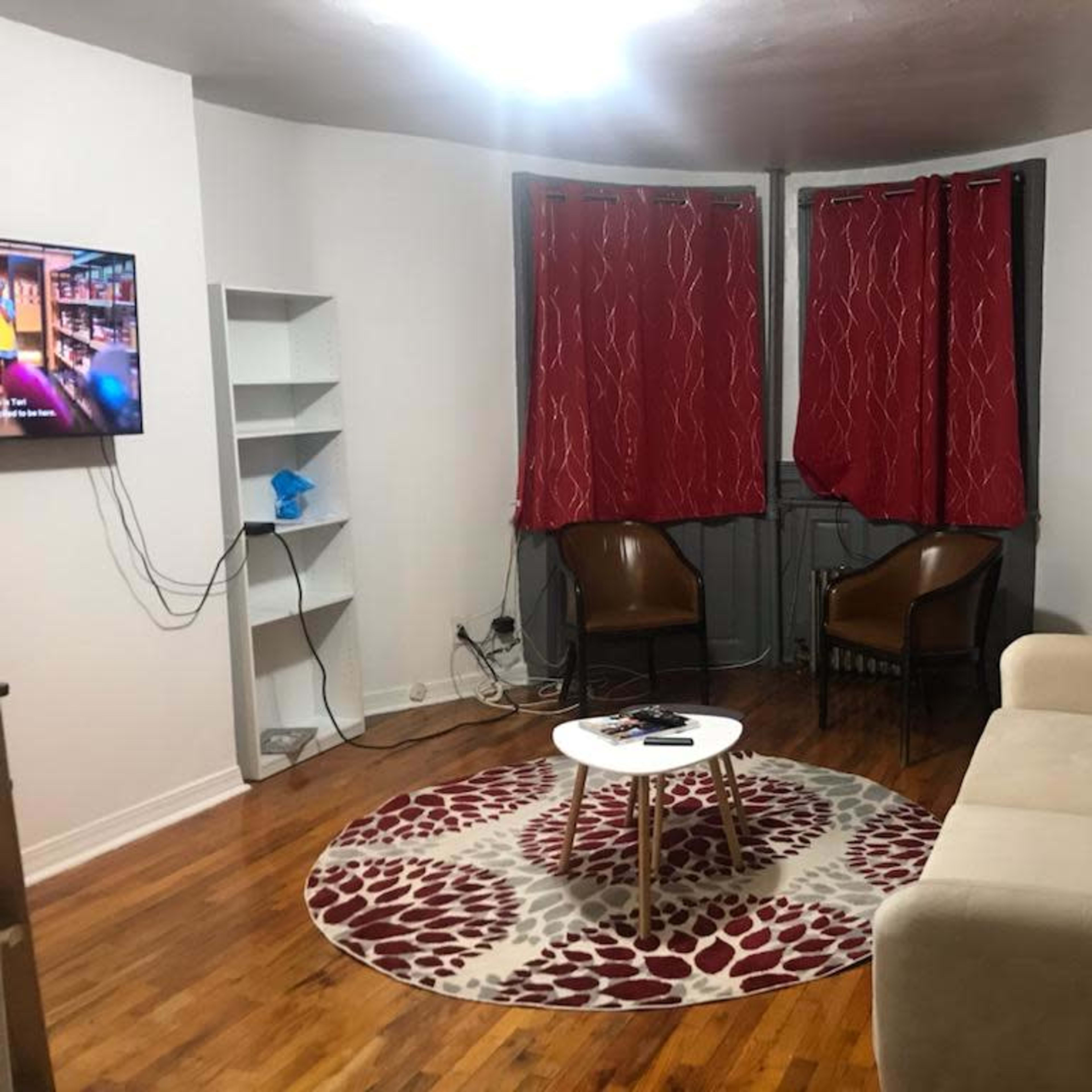 A small living room with a beige sofa, two brown chairs, a round coffee table, a television on the wall, and red curtains covering the window.