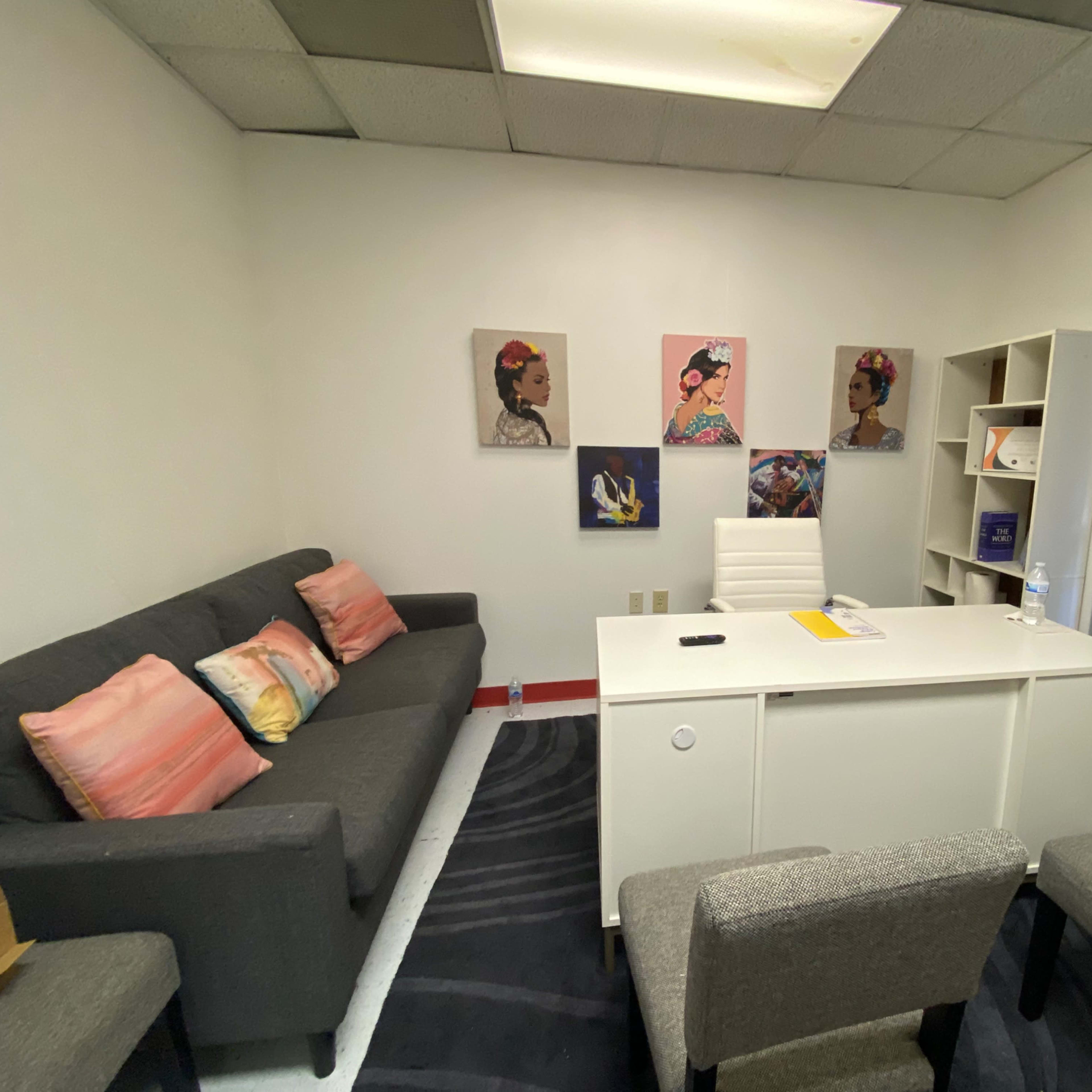 A small office with a white desk, a gray sofa with colorful pillows, and wall art featuring portraits.