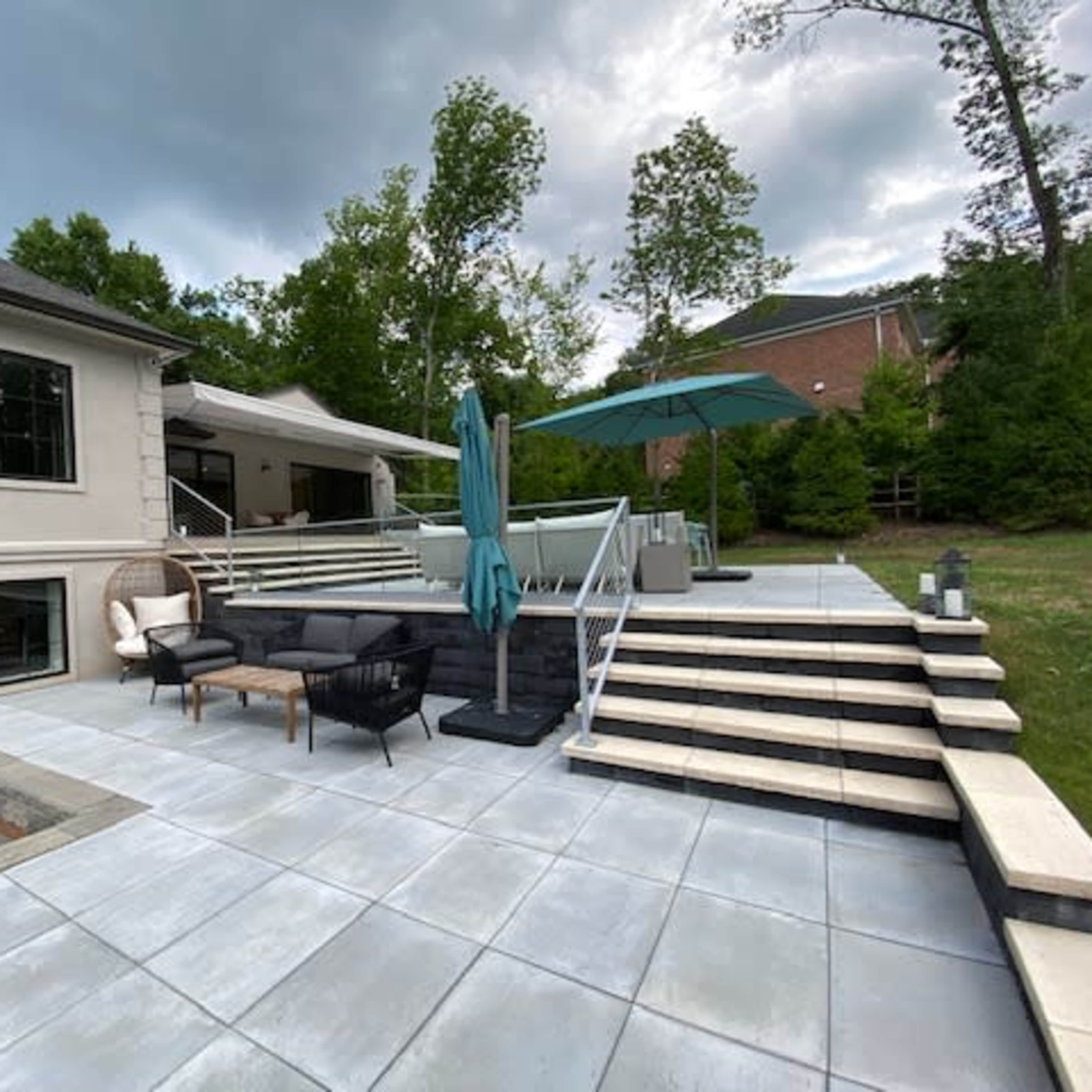 A modern patio area with outdoor furniture, a large umbrella, and stairs leading up to a house surrounded by greenery.