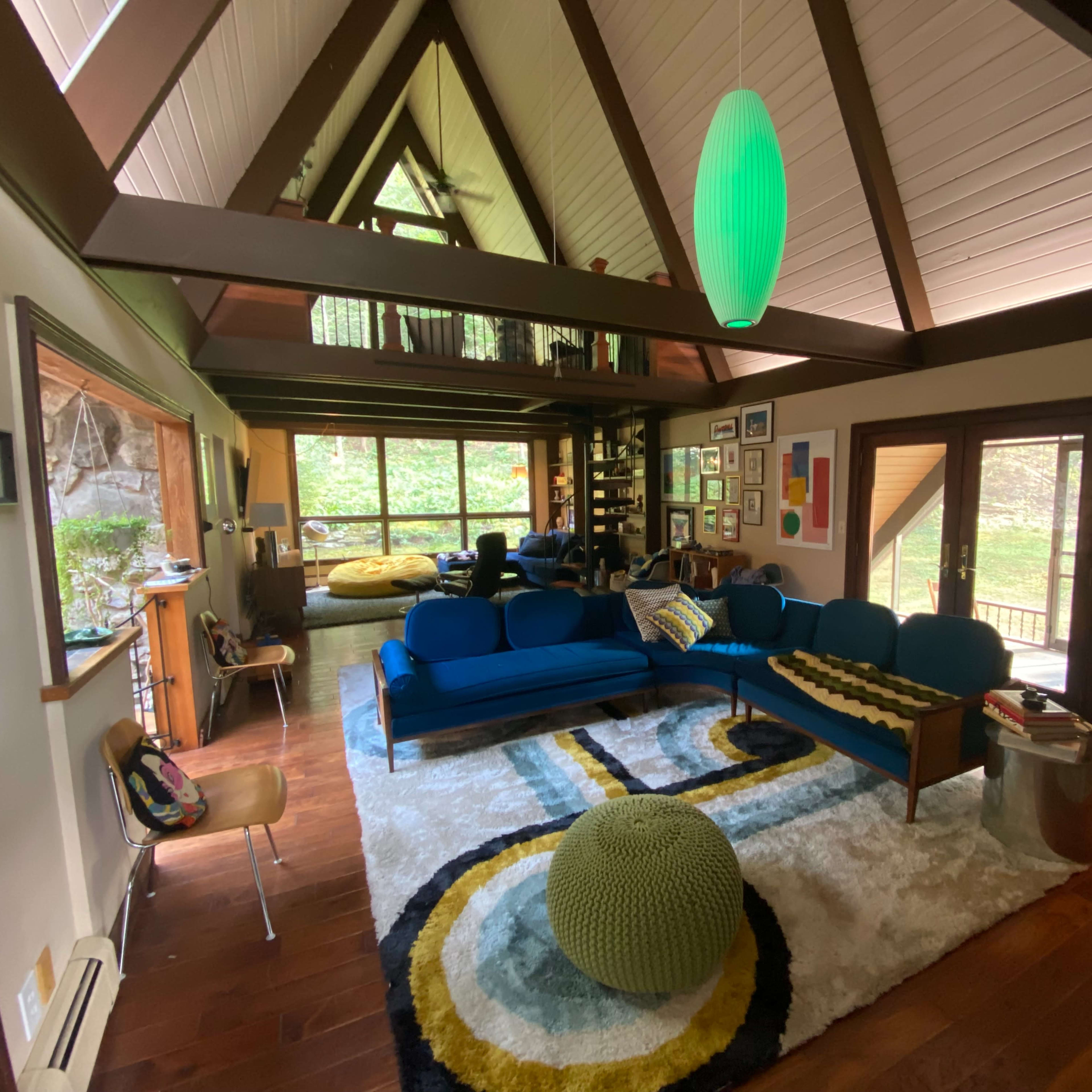 A cozy living room with a triangular wooden ceiling, blue sofas, a patterned rug, and large windows providing views of a green outdoor space.