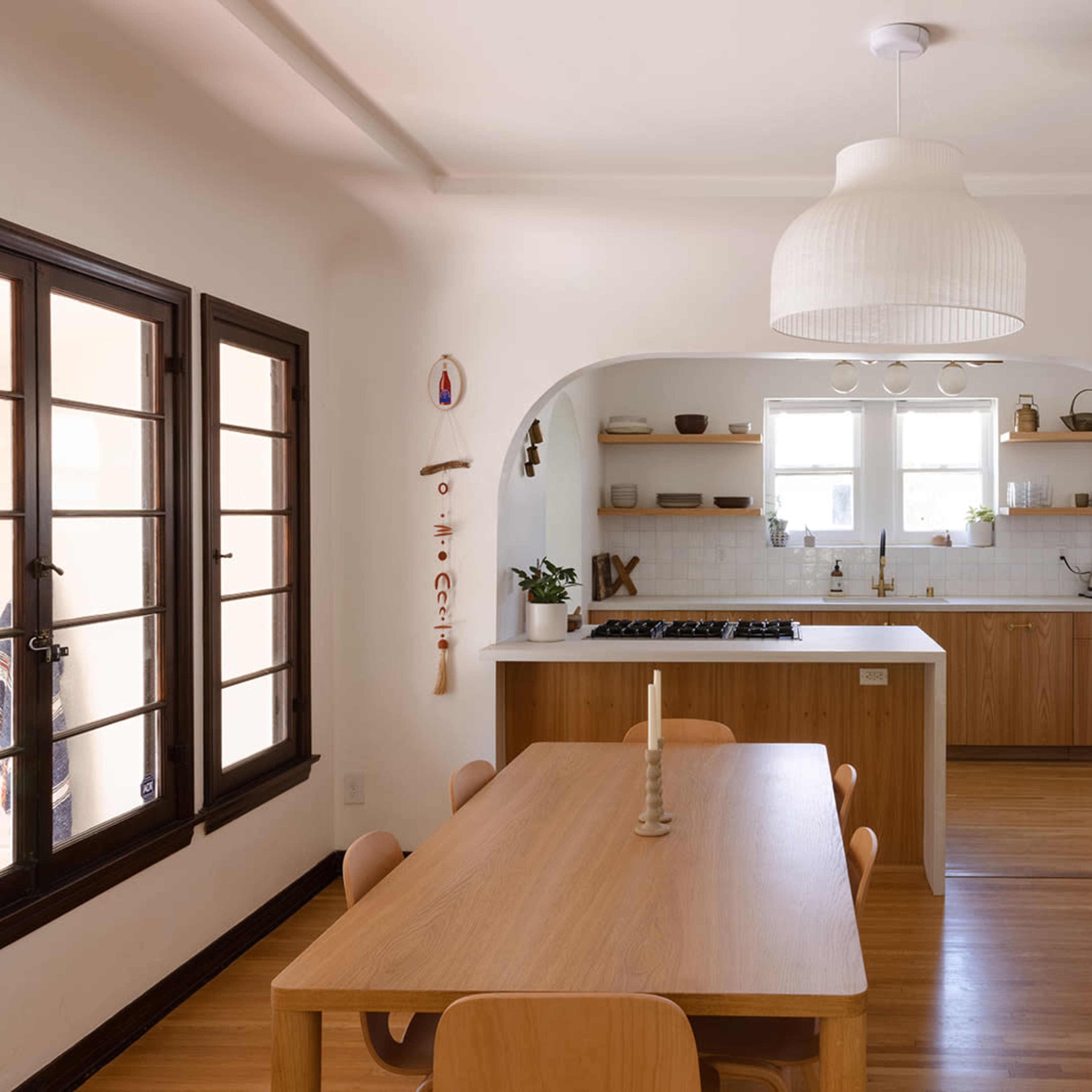 A modern dining area connected to a kitchen, featuring a wooden table with chairs, an archway leading to the kitchen, and shelves displaying kitchenware.