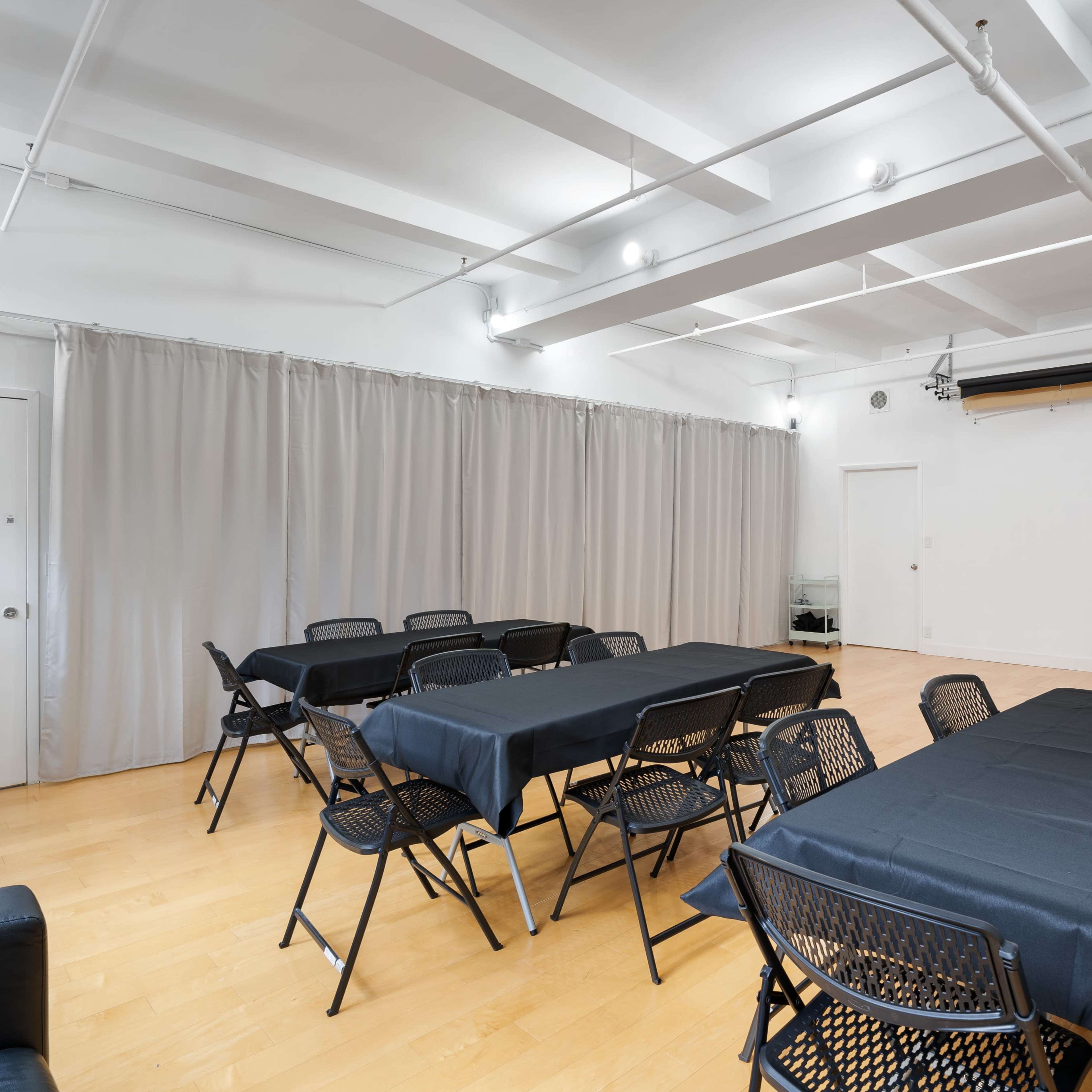 A spacious room is set up with black tables and chairs, divided by gray curtains, and features a plain wall at one end.