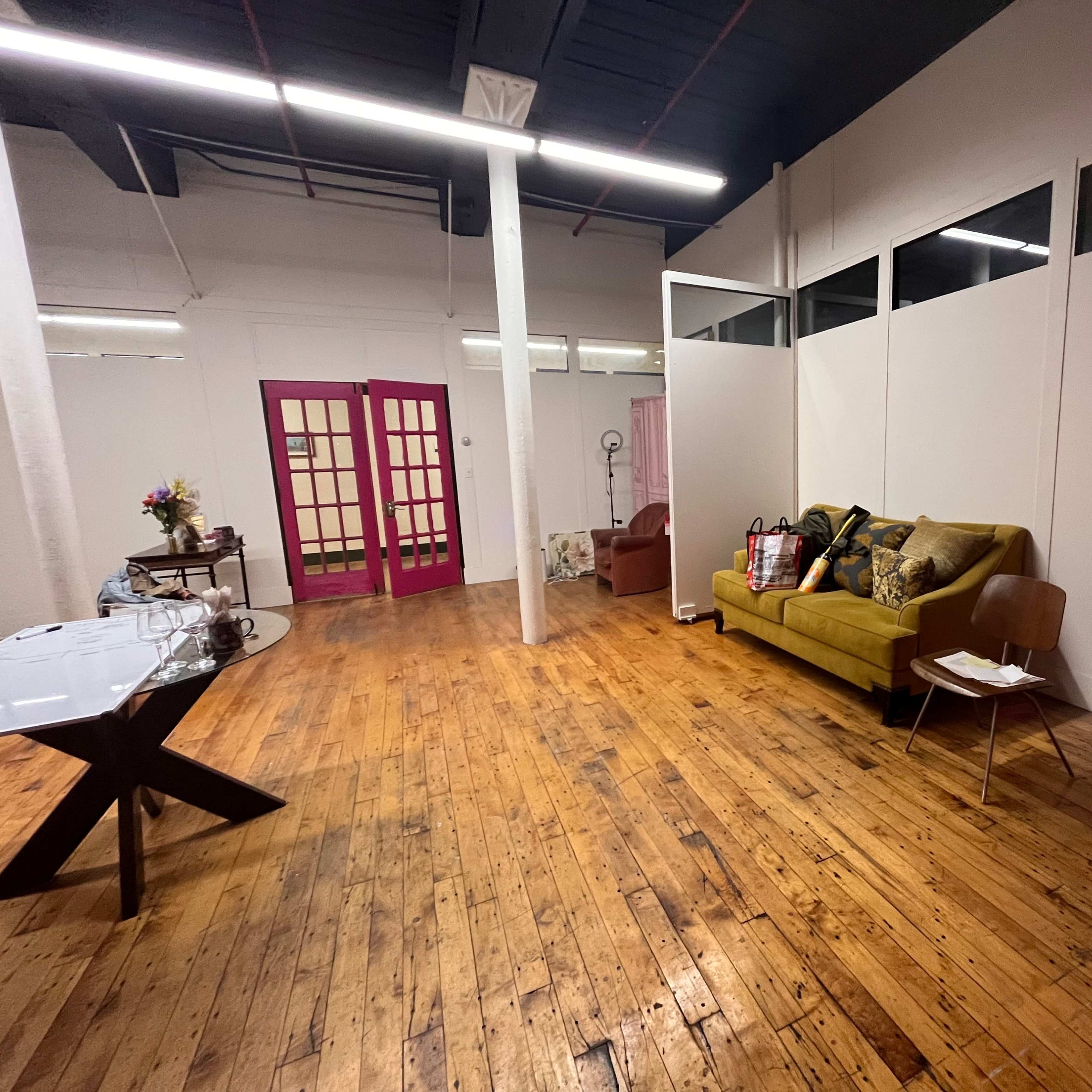 A spacious room with wooden floors, a yellow couch, a small chair, a table, and a sectioned-off area with a pink door at one end.