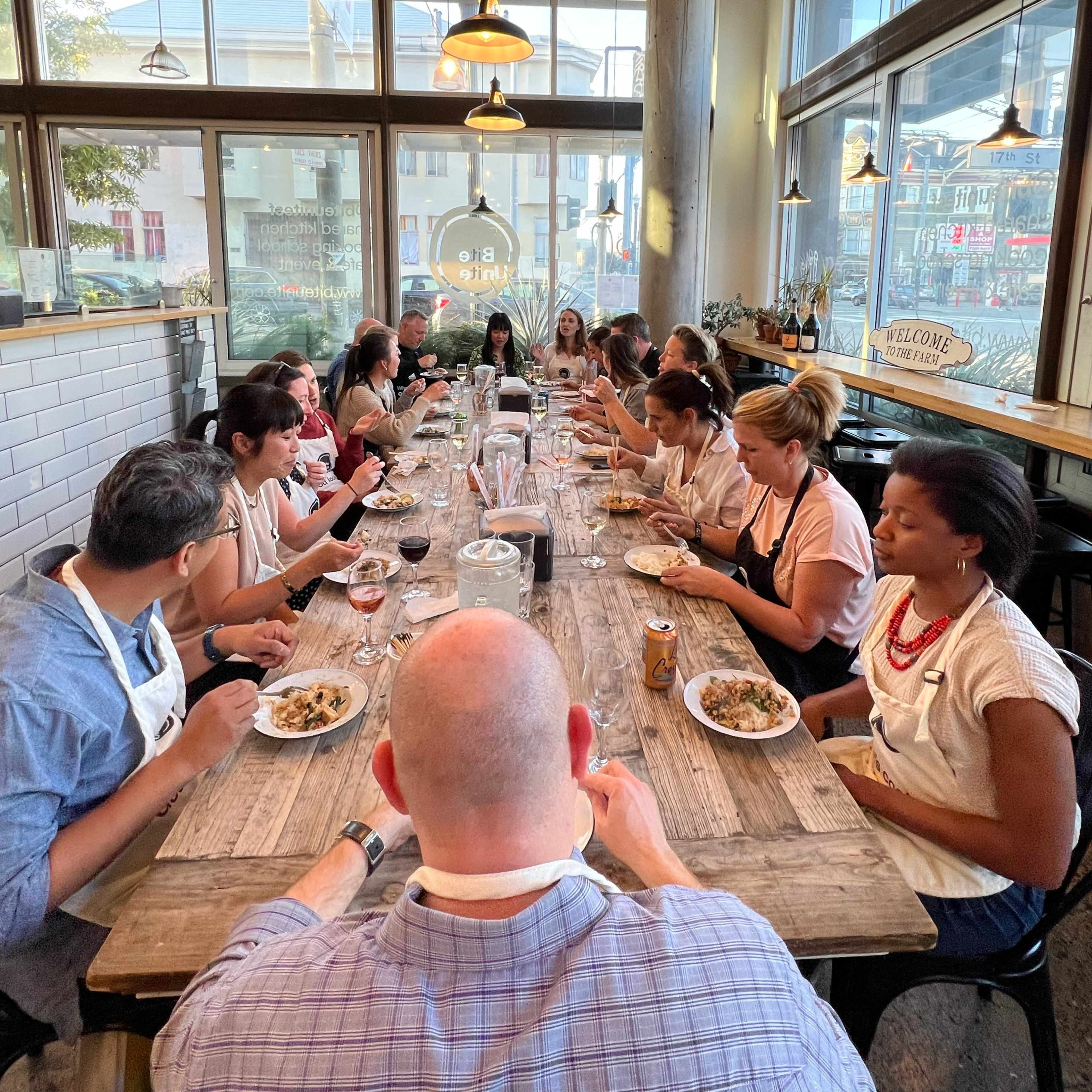 A group of people sits at a long wooden table, enjoying a meal together in a brightly lit restaurant with large windows.