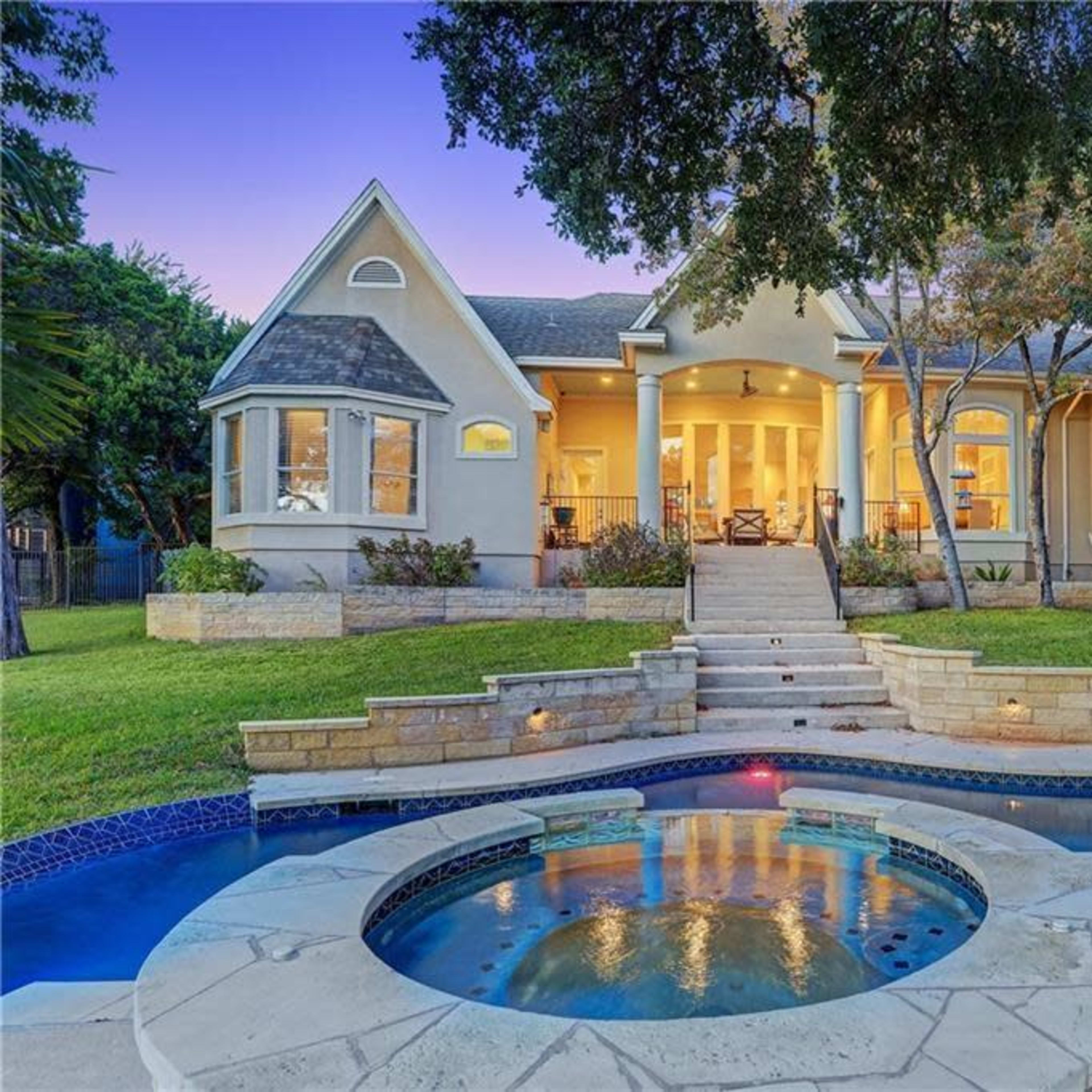 A two-story house with large windows and a stone pathway leads to a landscaped yard featuring a pool and a hot tub.