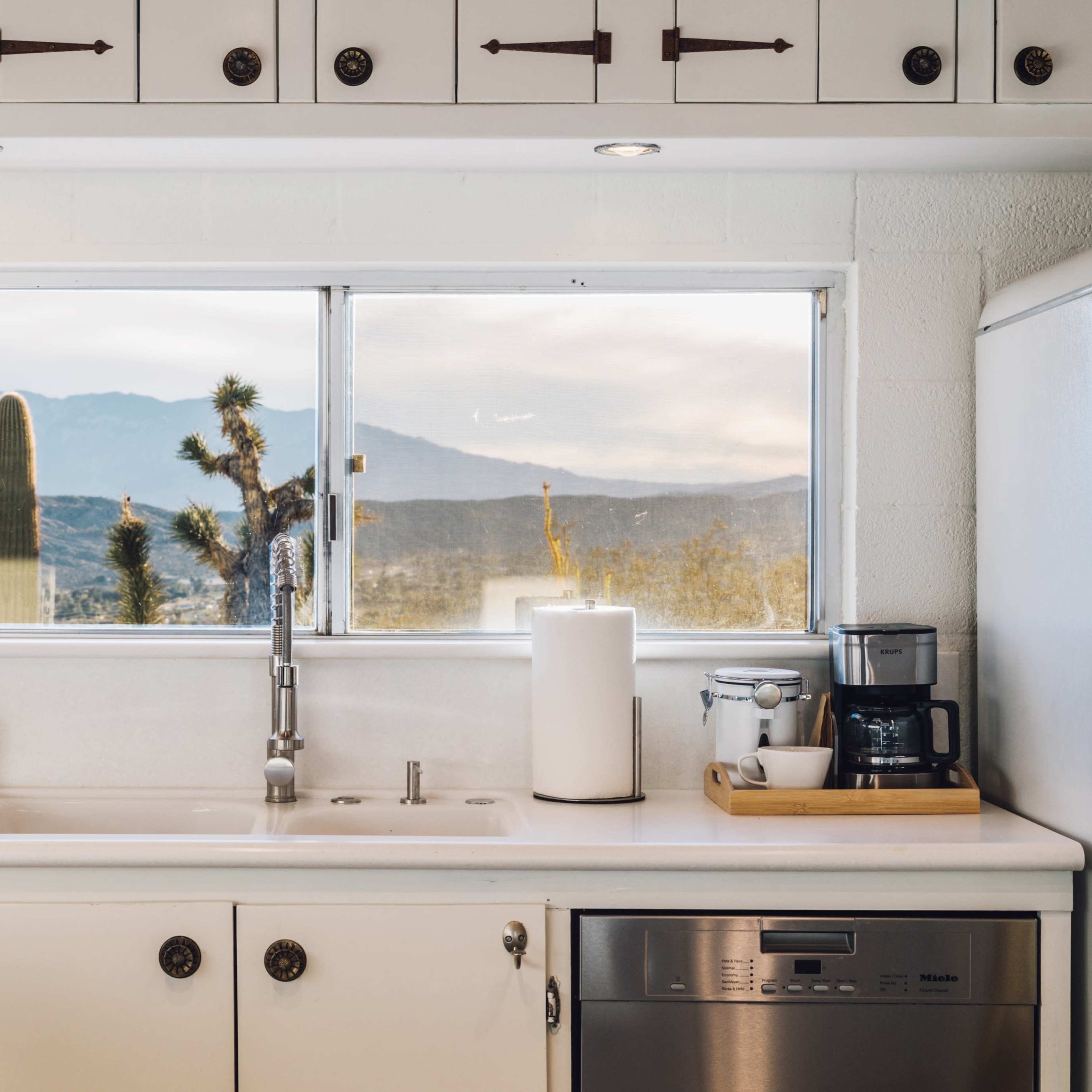 A modern kitchen features white cabinets, a sink beneath a large window, and a view of cacti and mountains outside.