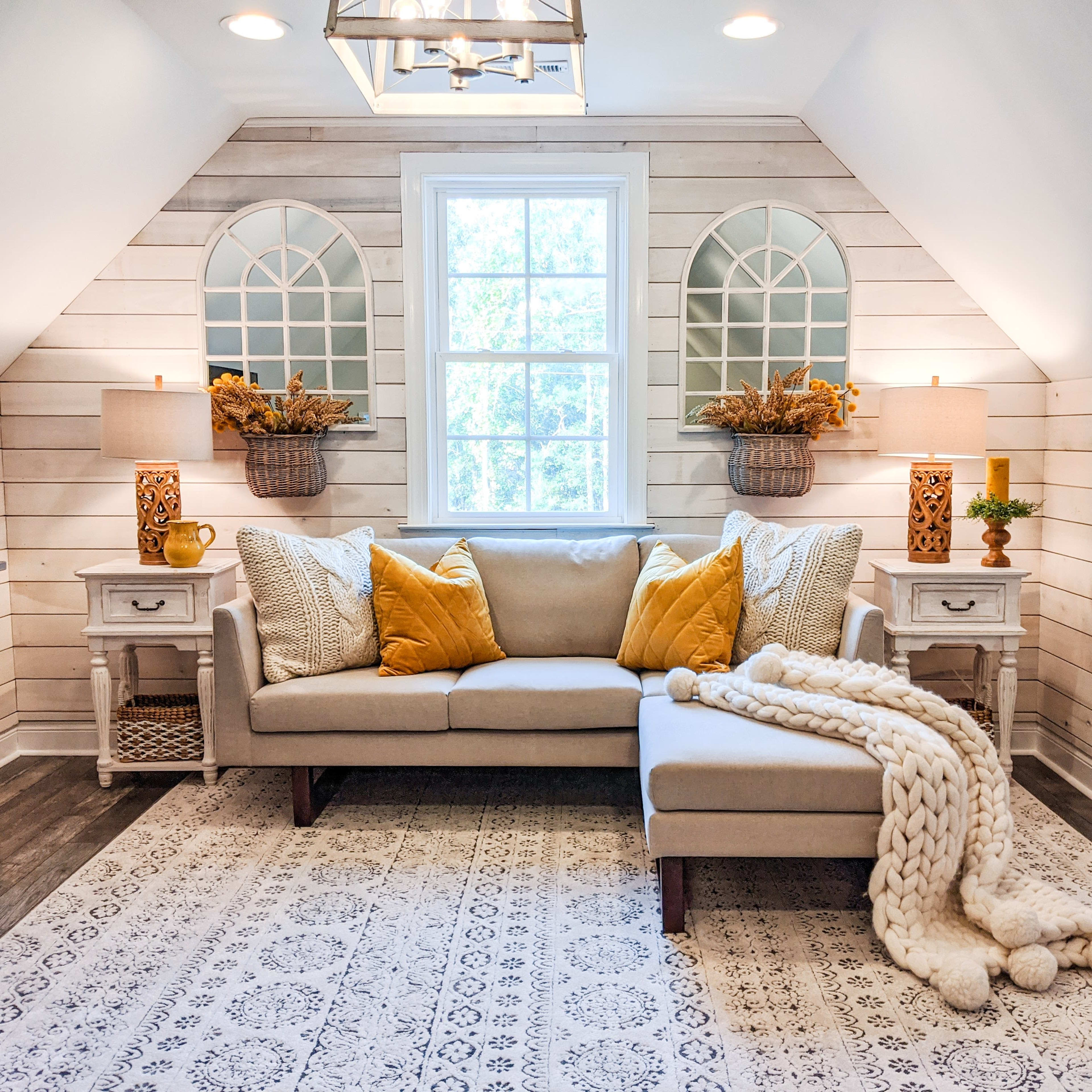 A cozy, well-decorated attic space featuring a light-colored sofa, decorative pillows, a knitted blanket, and two large windows with natural light.