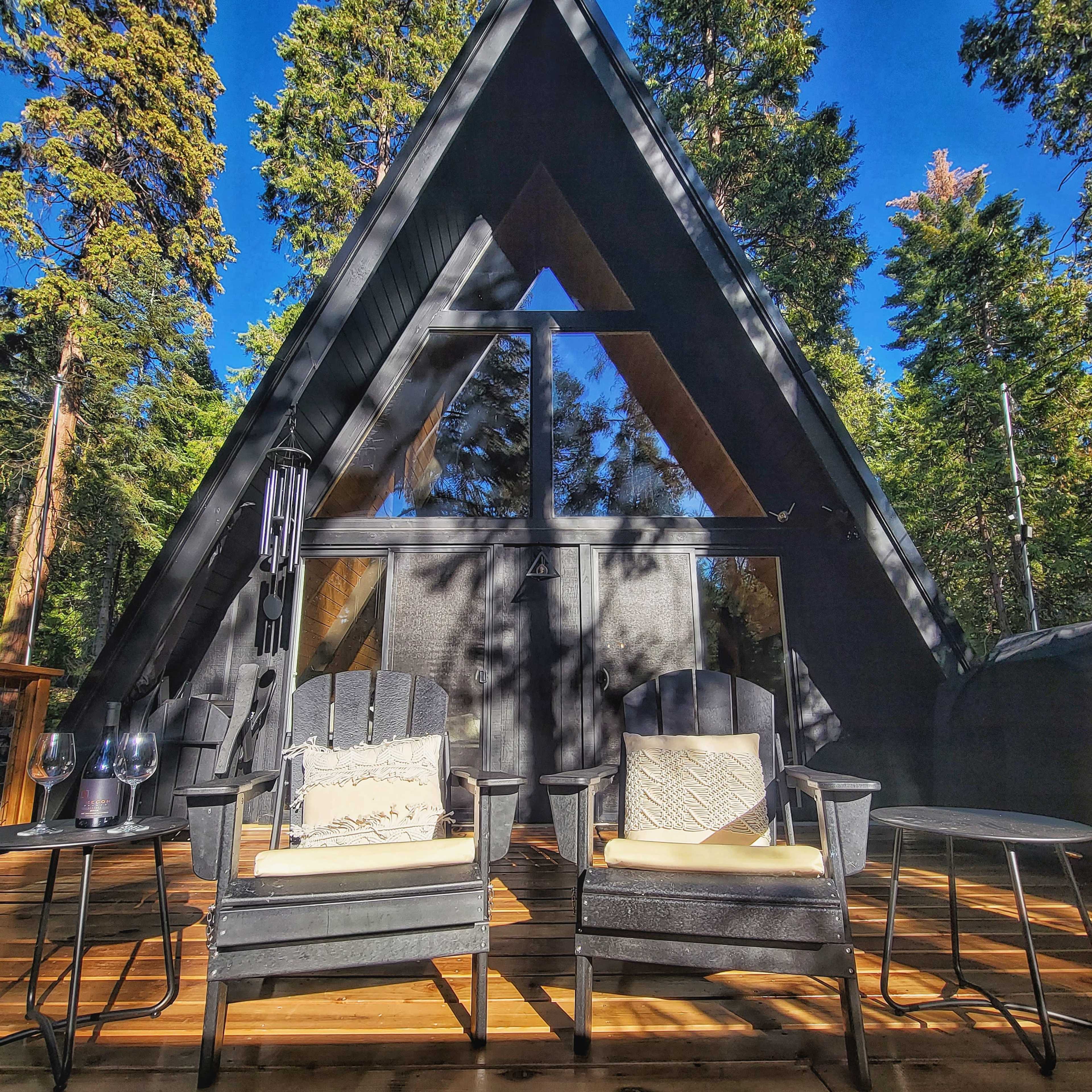 A modern A-frame cabin surrounded by tall trees, with two black chairs and a small table on a wooden deck in front.