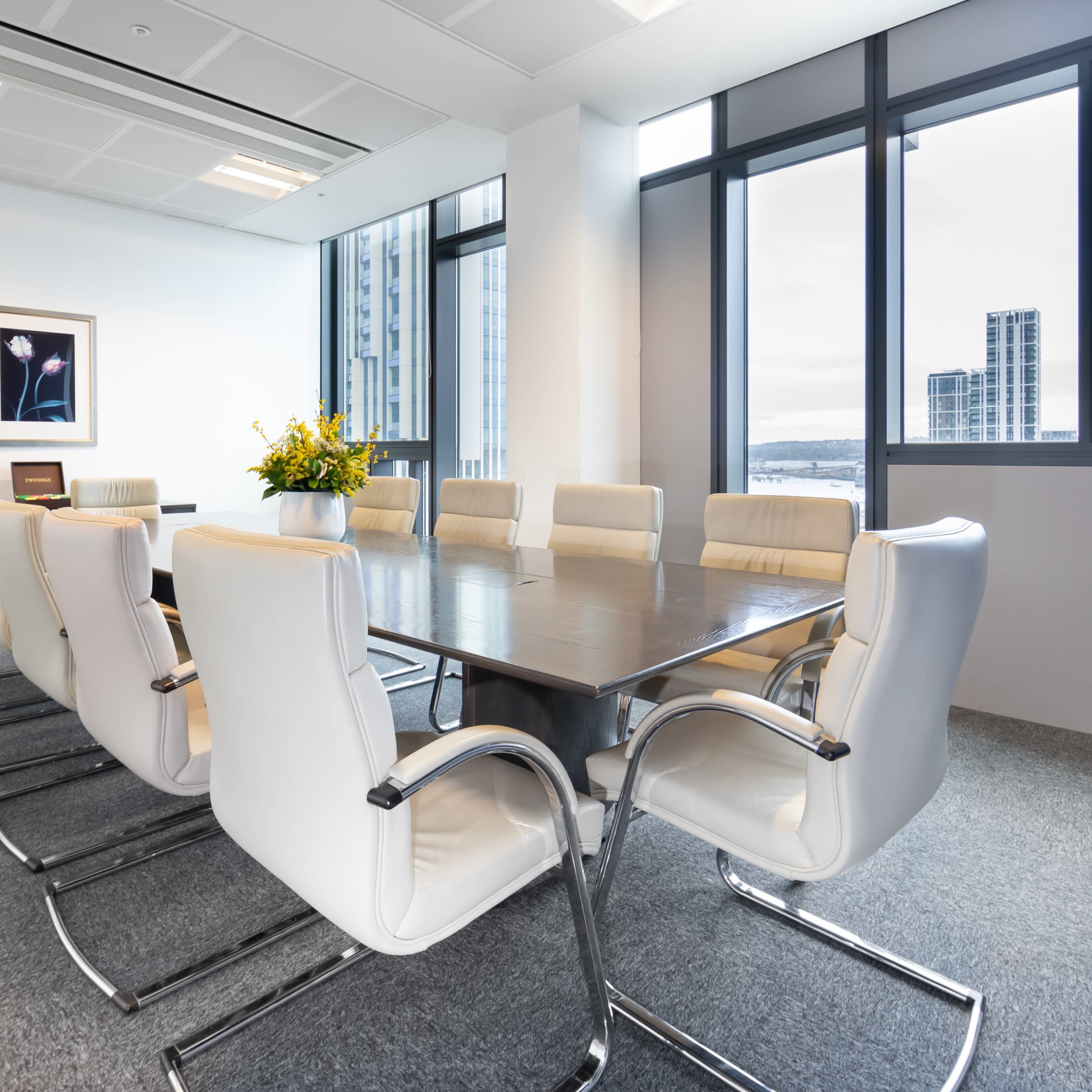 A modern conference room features a long wooden table surrounded by twelve white chairs, with large windows offering a view of the city.