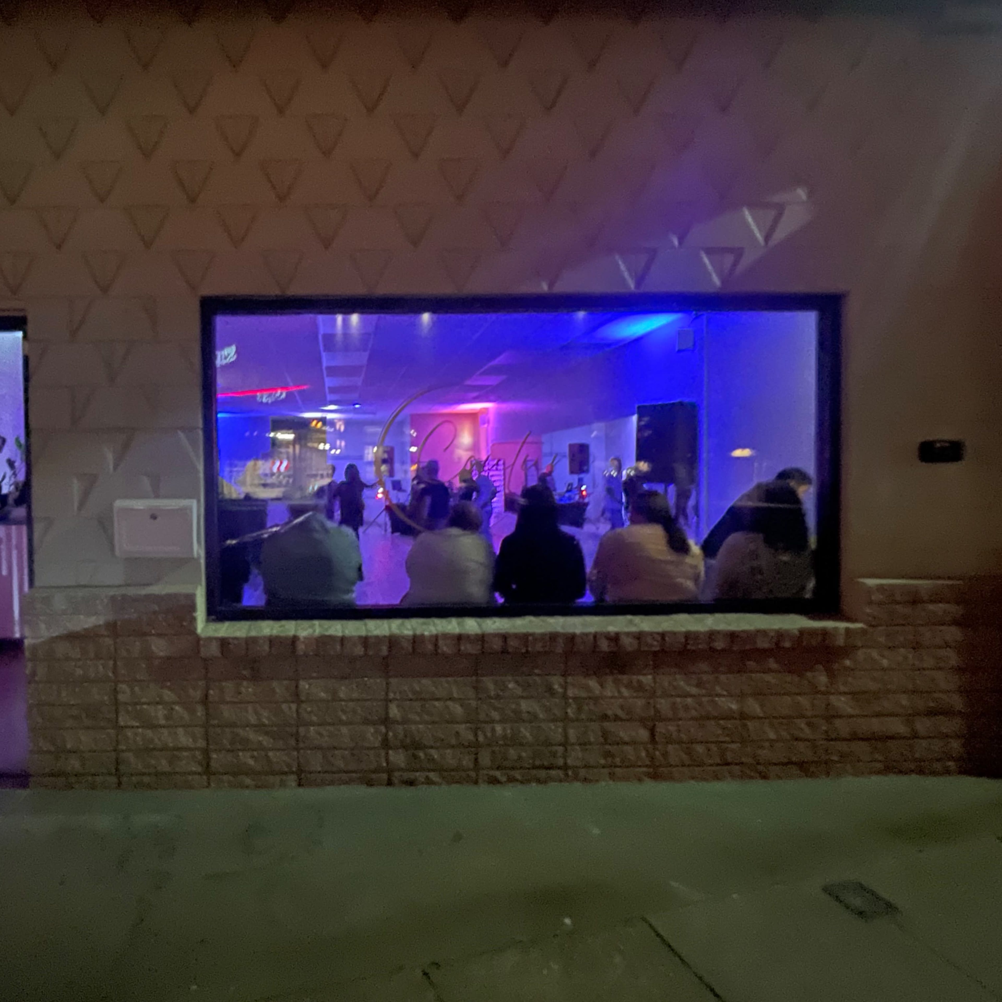 A group of people is seated in front of a brightly lit stage inside a venue, visible through a large window.