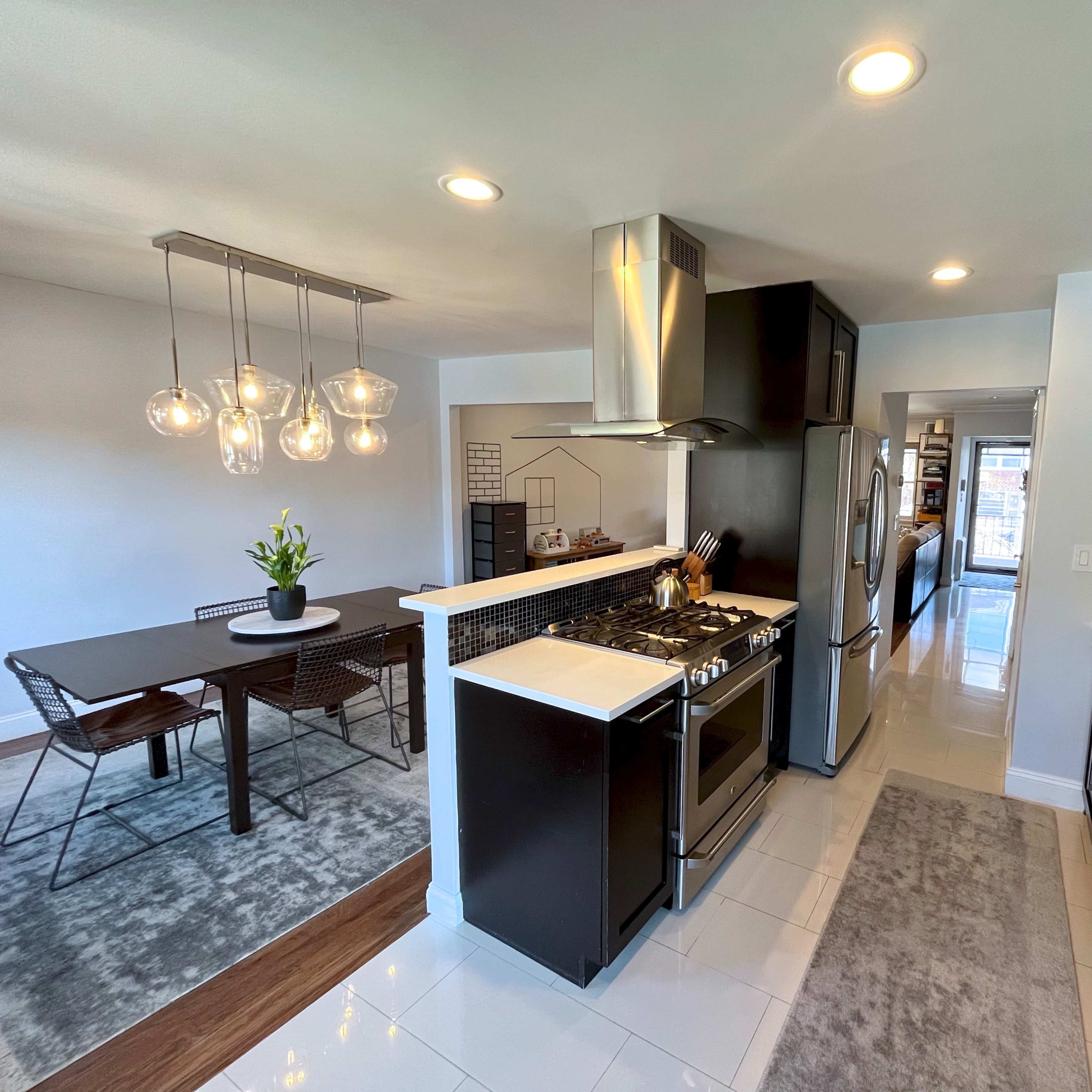 A modern kitchen with dark cabinetry, a stainless steel range, and a dining area with a table and chairs illuminated by pendant lights.