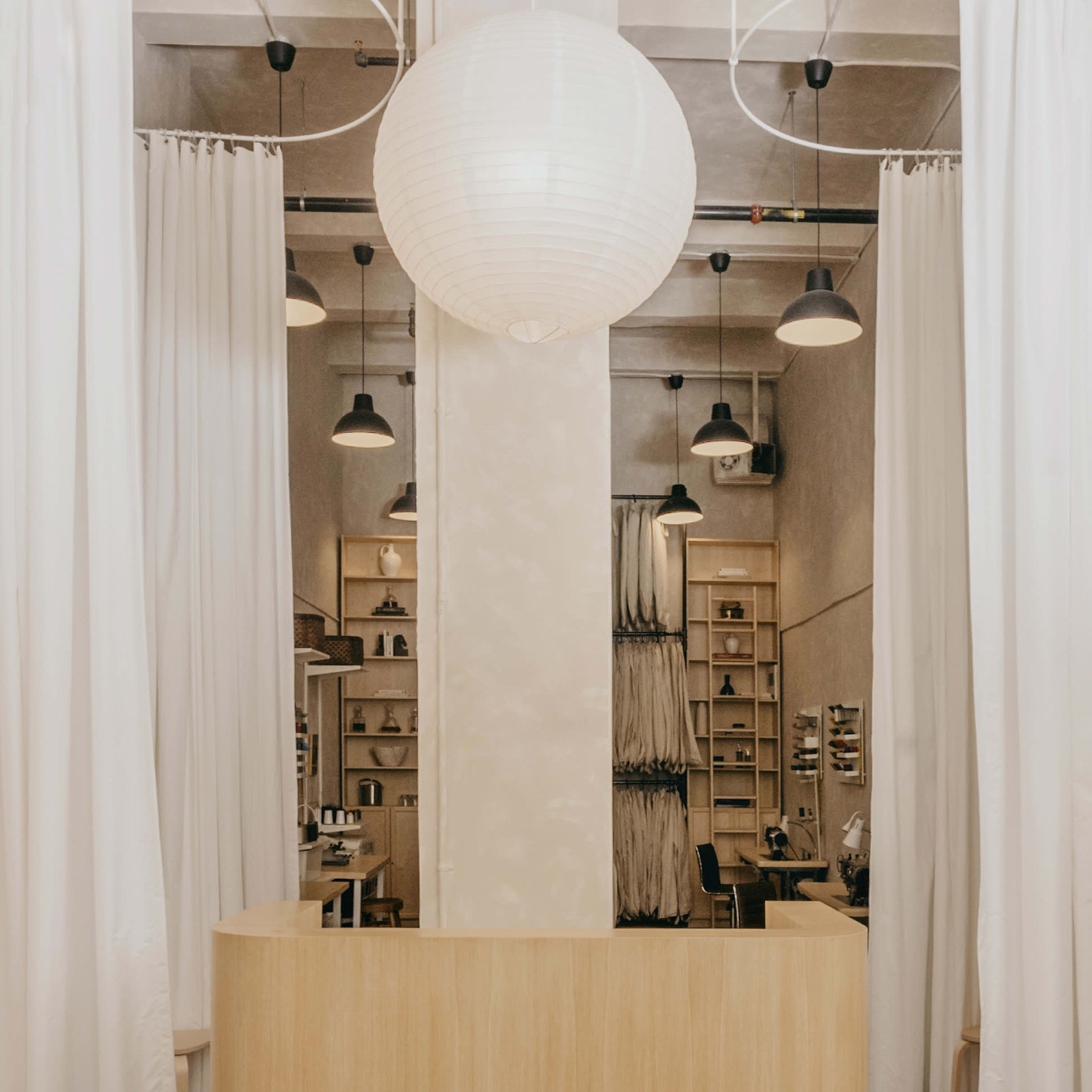 A wooden reception desk stands in a minimalist space with white curtains and a large paper lantern overhead.