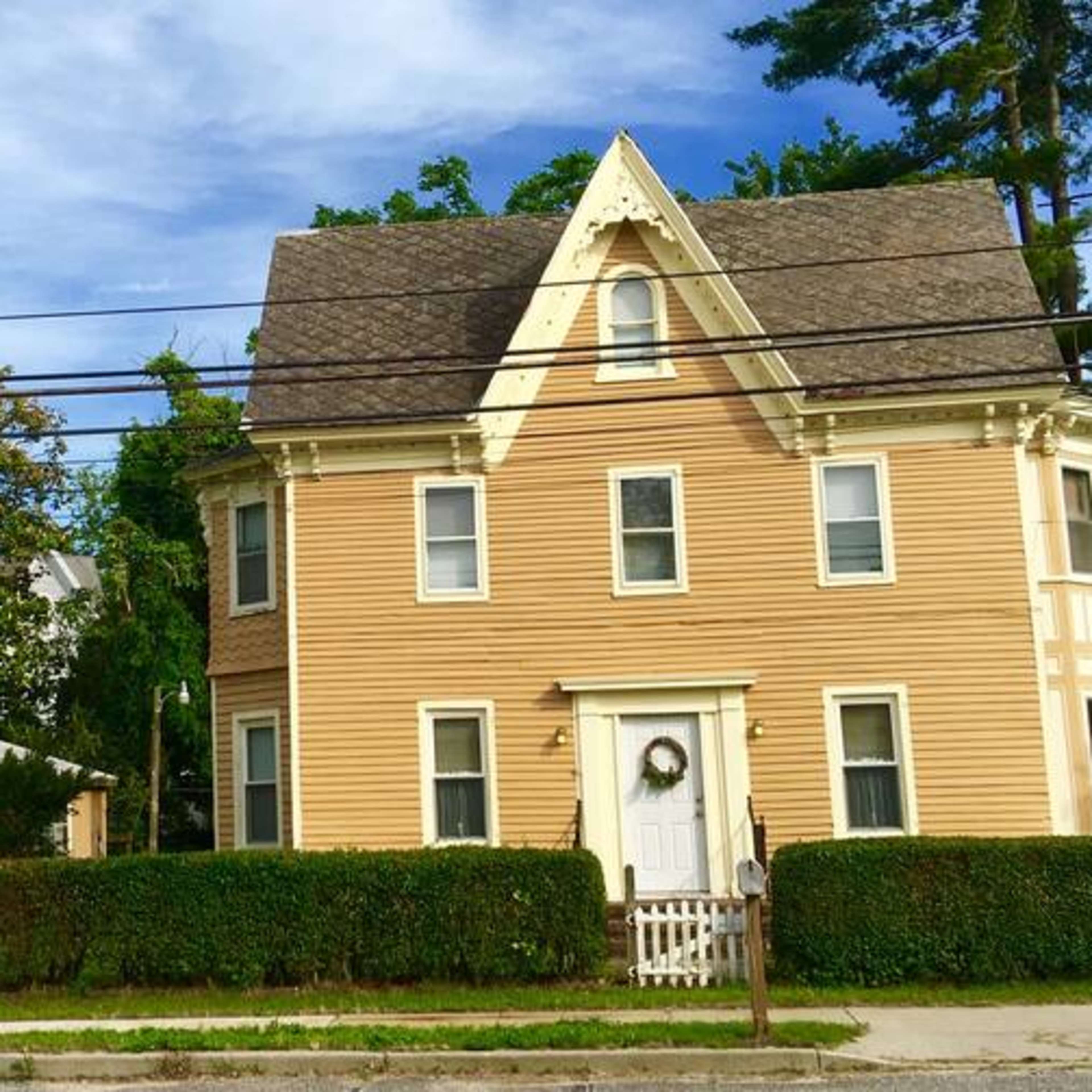 A two-story yellow house with a gabled roof next to a smaller yellow barn, surrounded by a neatly trimmed hedge and lined by power lines.