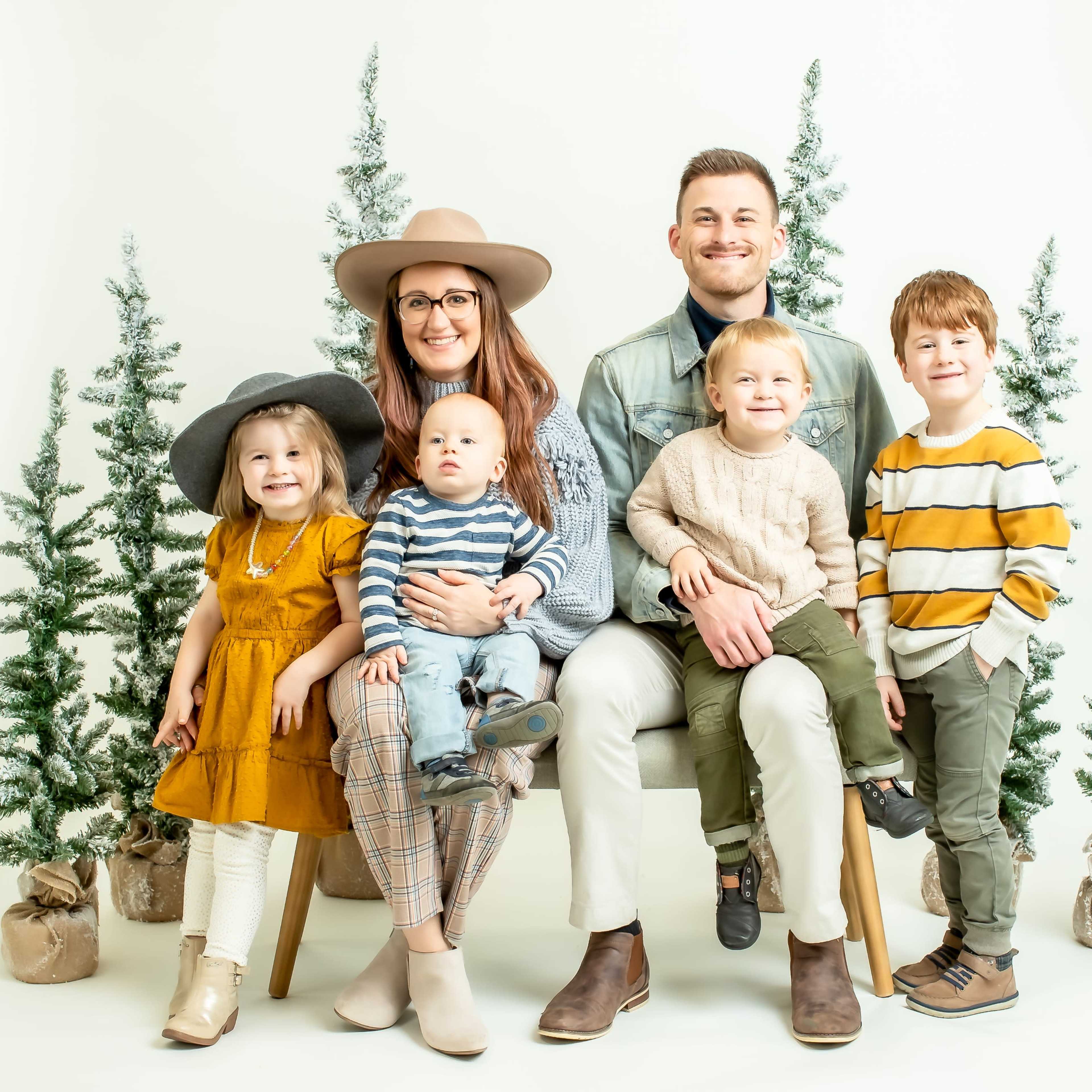 A family of five, including three children, poses together on a chair in front of small decorated evergreen trees.