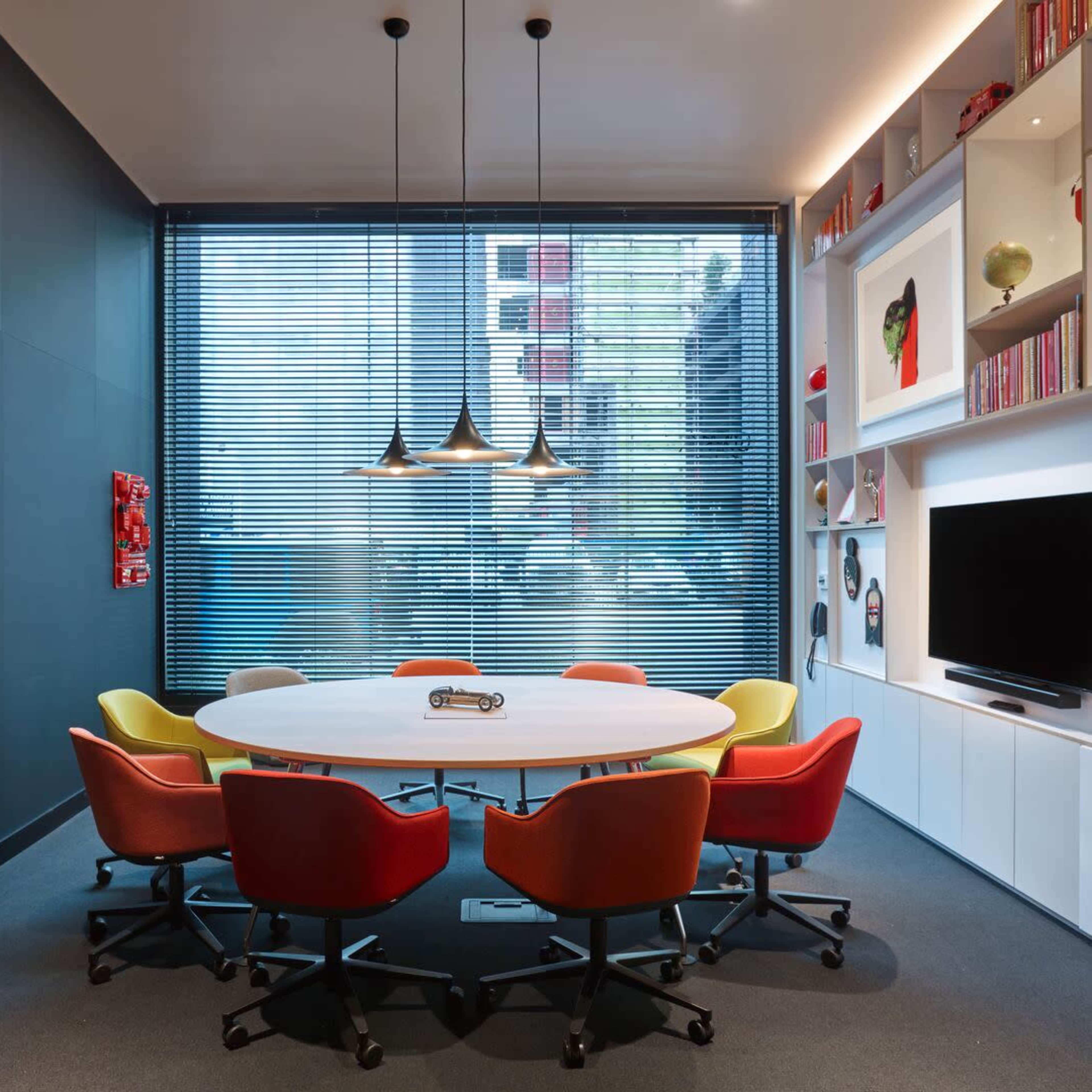 A brightly lit conference room features a large oval table surrounded by eight colorful chairs, with a window displaying an urban view and shelves filled with decorative items and books.