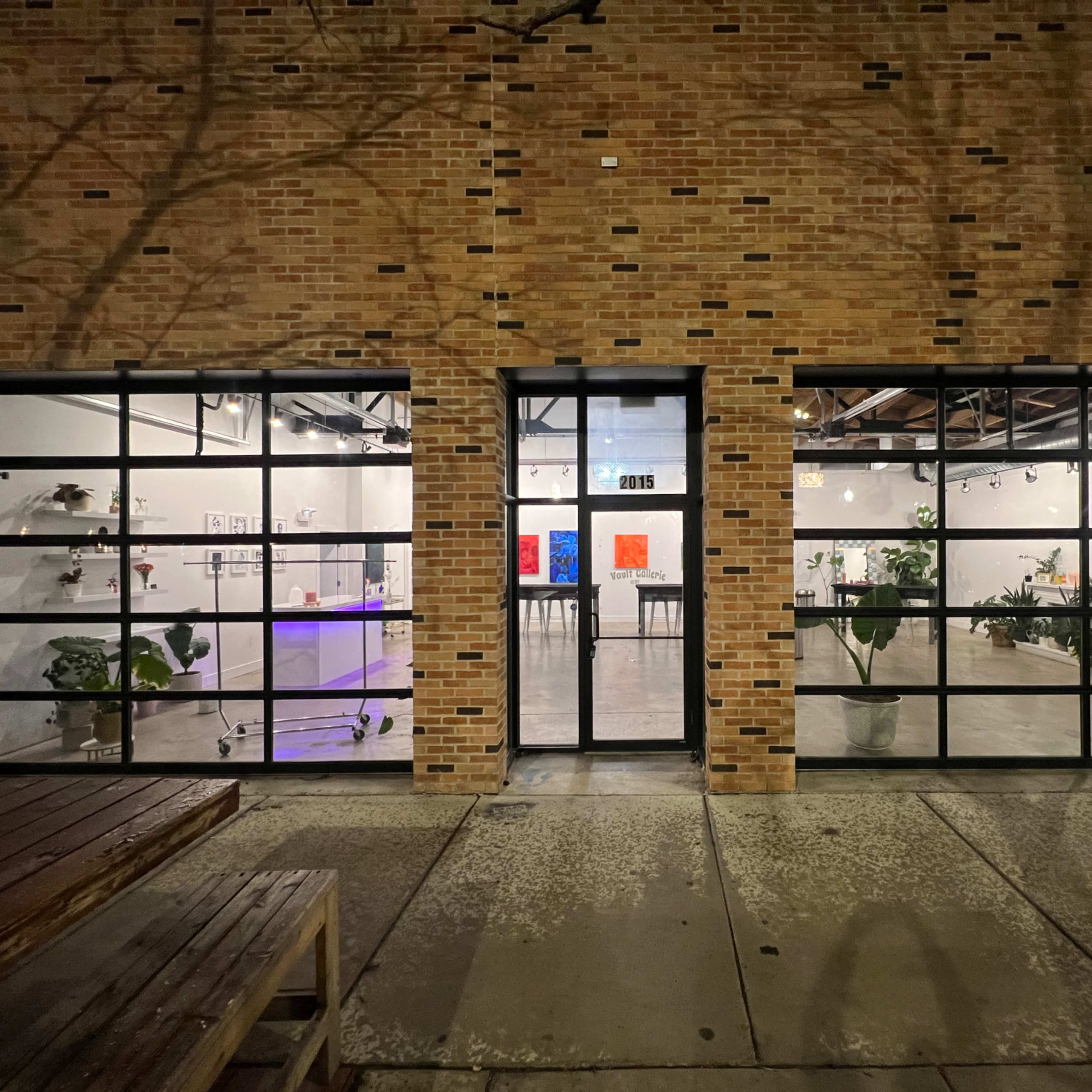 A brick storefront with large, black-framed windows revealing an art gallery interior featuring various artworks and plants.