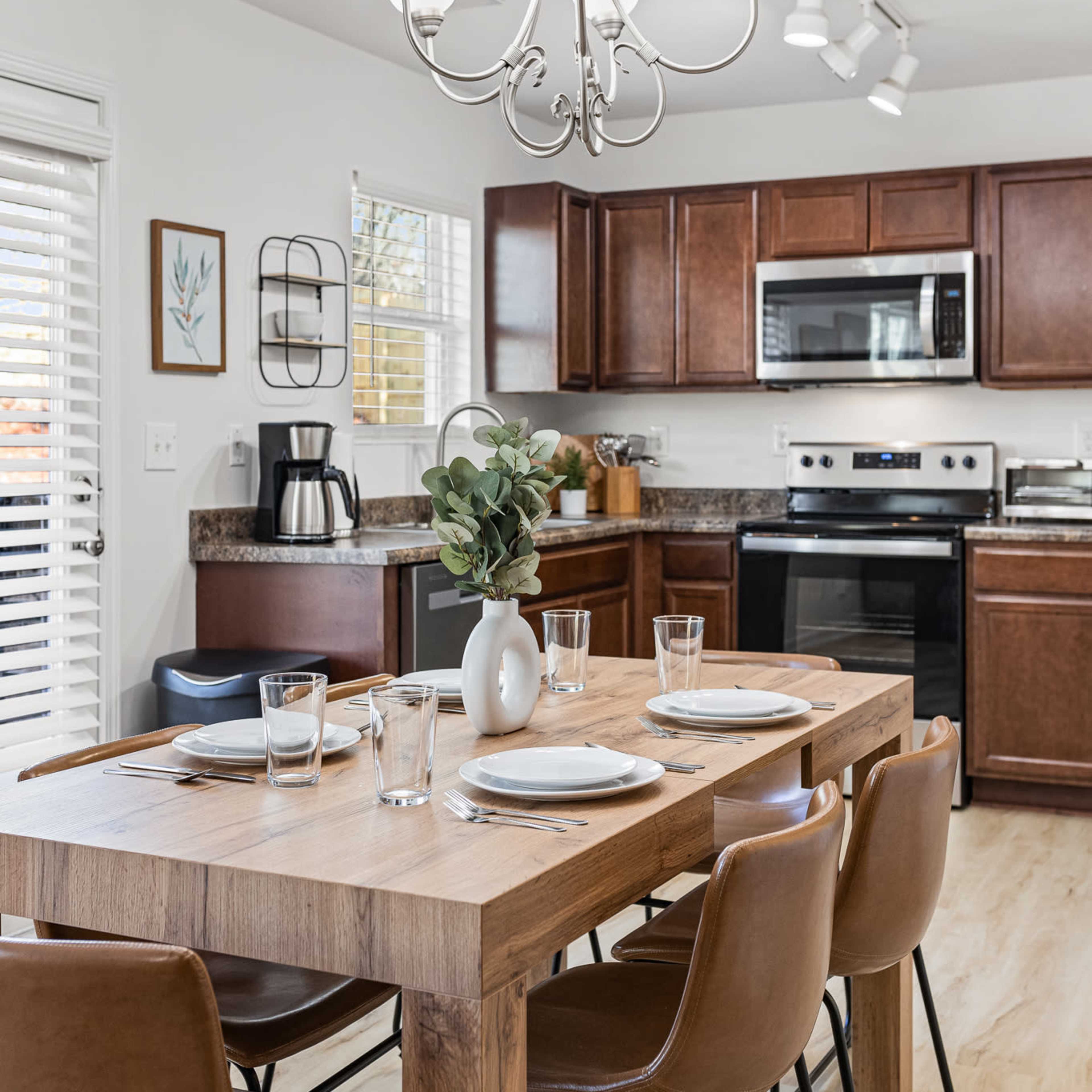 A modern kitchen features a wooden dining table set for four, surrounded by brown chairs, with cabinets and appliances in the background.