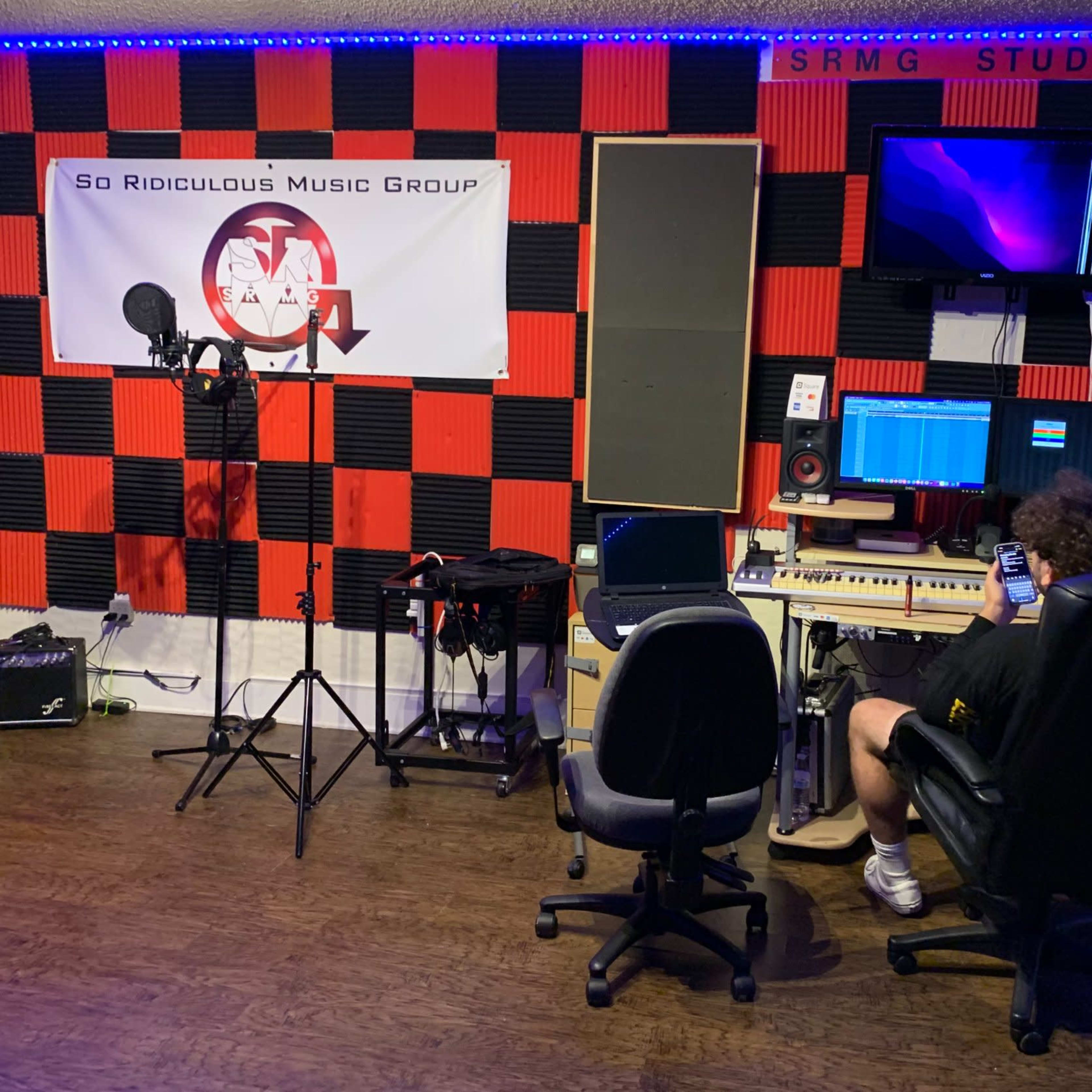 A person sits on a chair using a phone in a music studio with black and red soundproofing, microphones, and audio equipment.