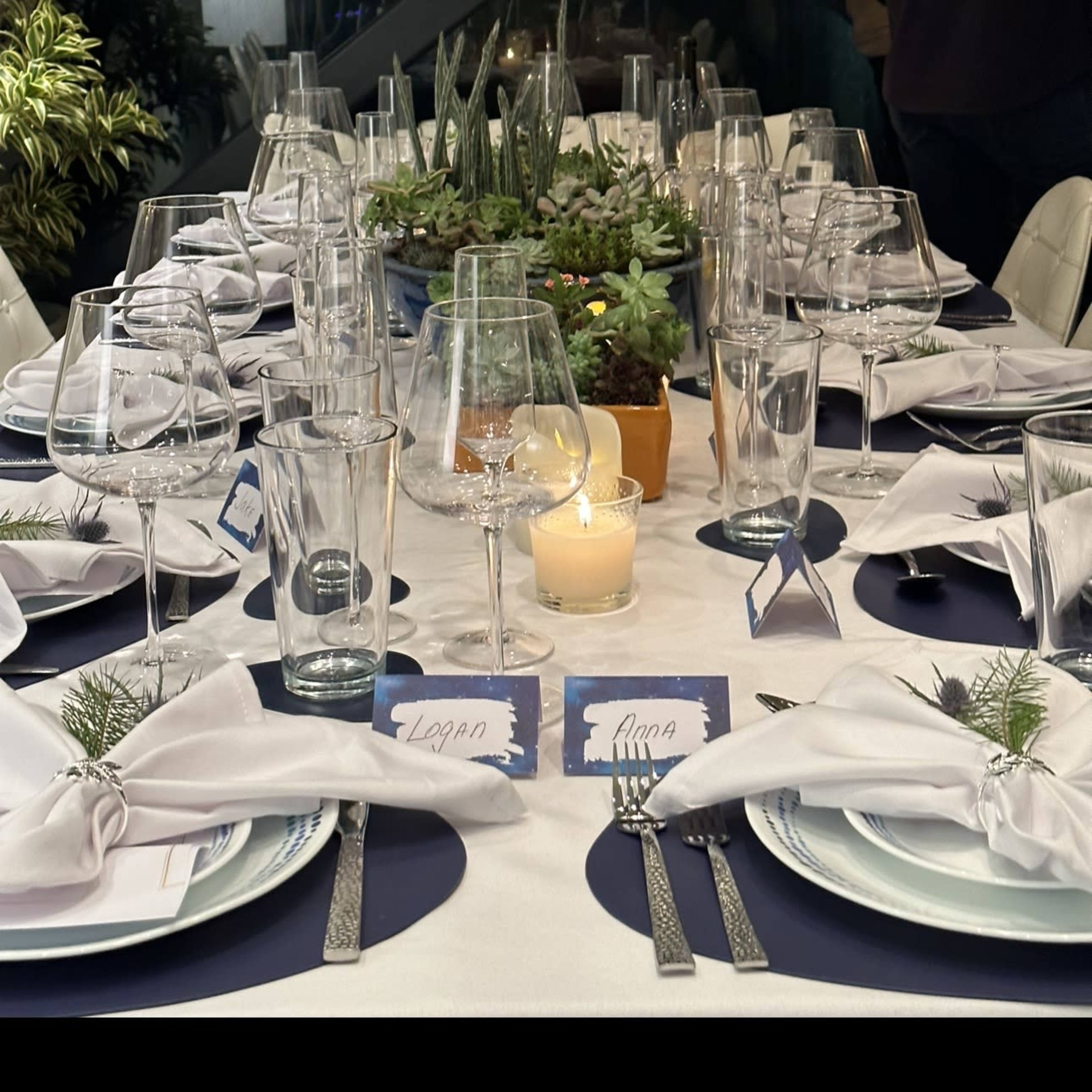 A long dining table is set with white tablecloths, glassware, silverware, and decorative greenery, with a single candle illuminating the center.