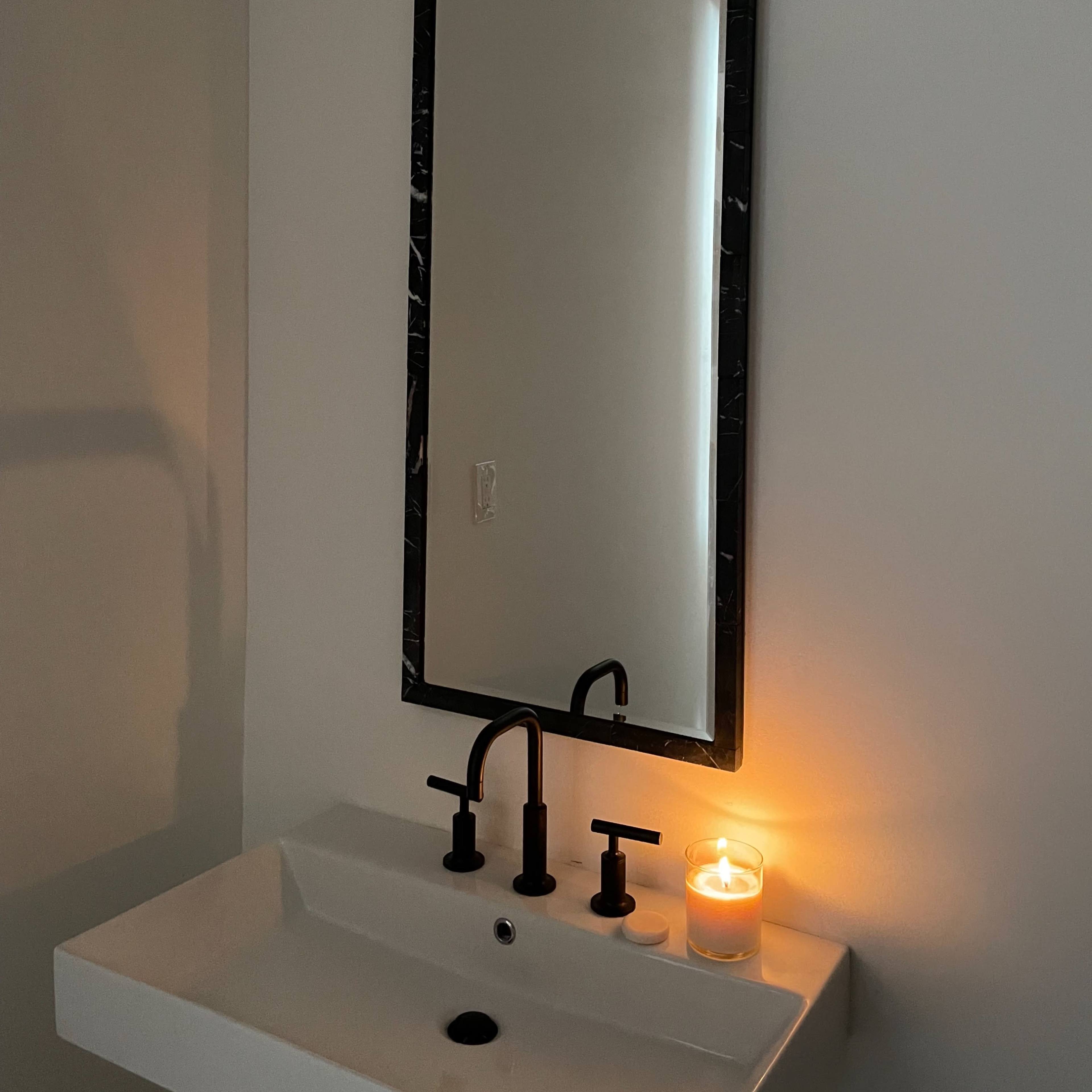 A bathroom sink with a black faucet and a lit candle sits next to a large rectangular mirror on the wall.