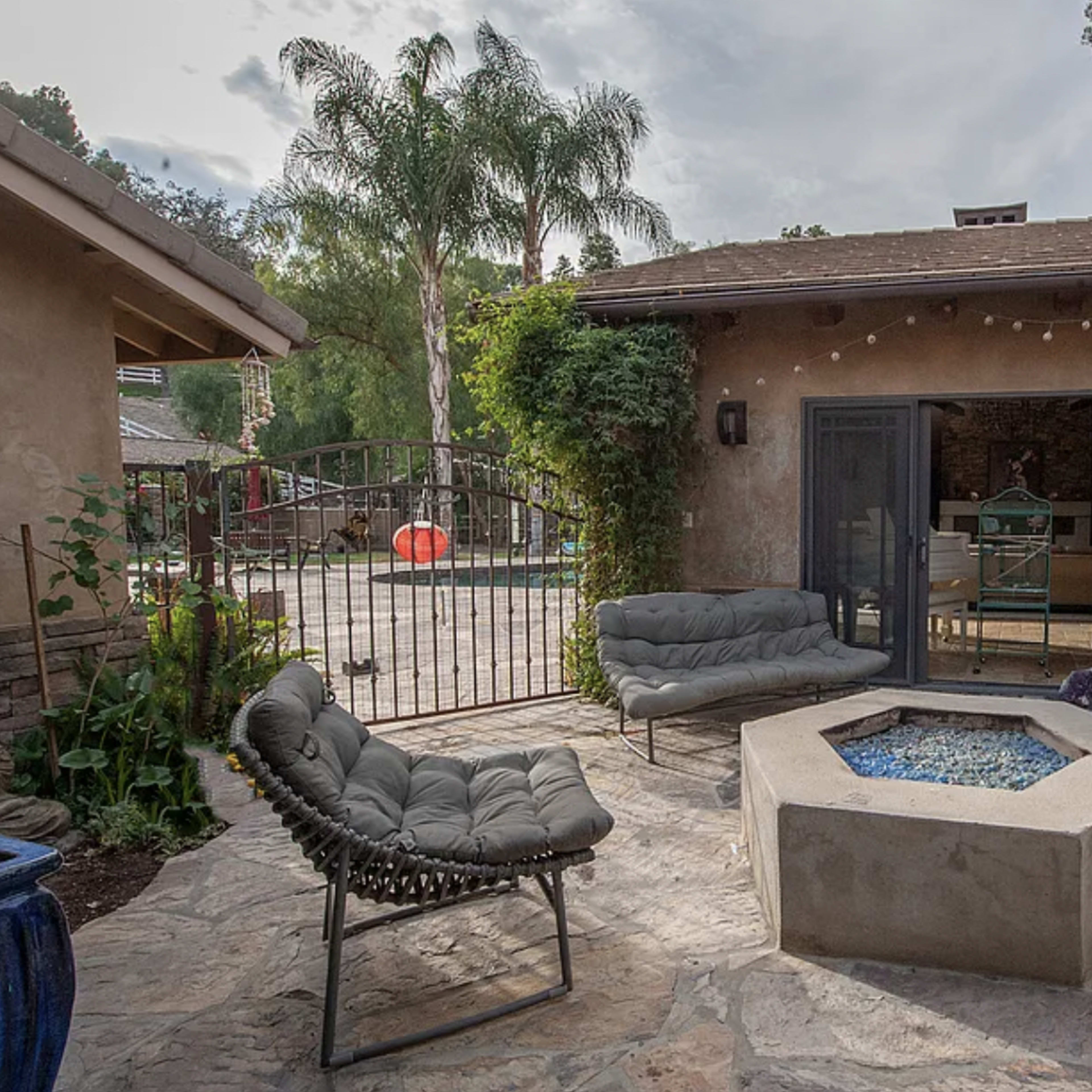 A landscaped patio features a central fire pit surrounded by modern seating and a gated entrance leading to a pool area.