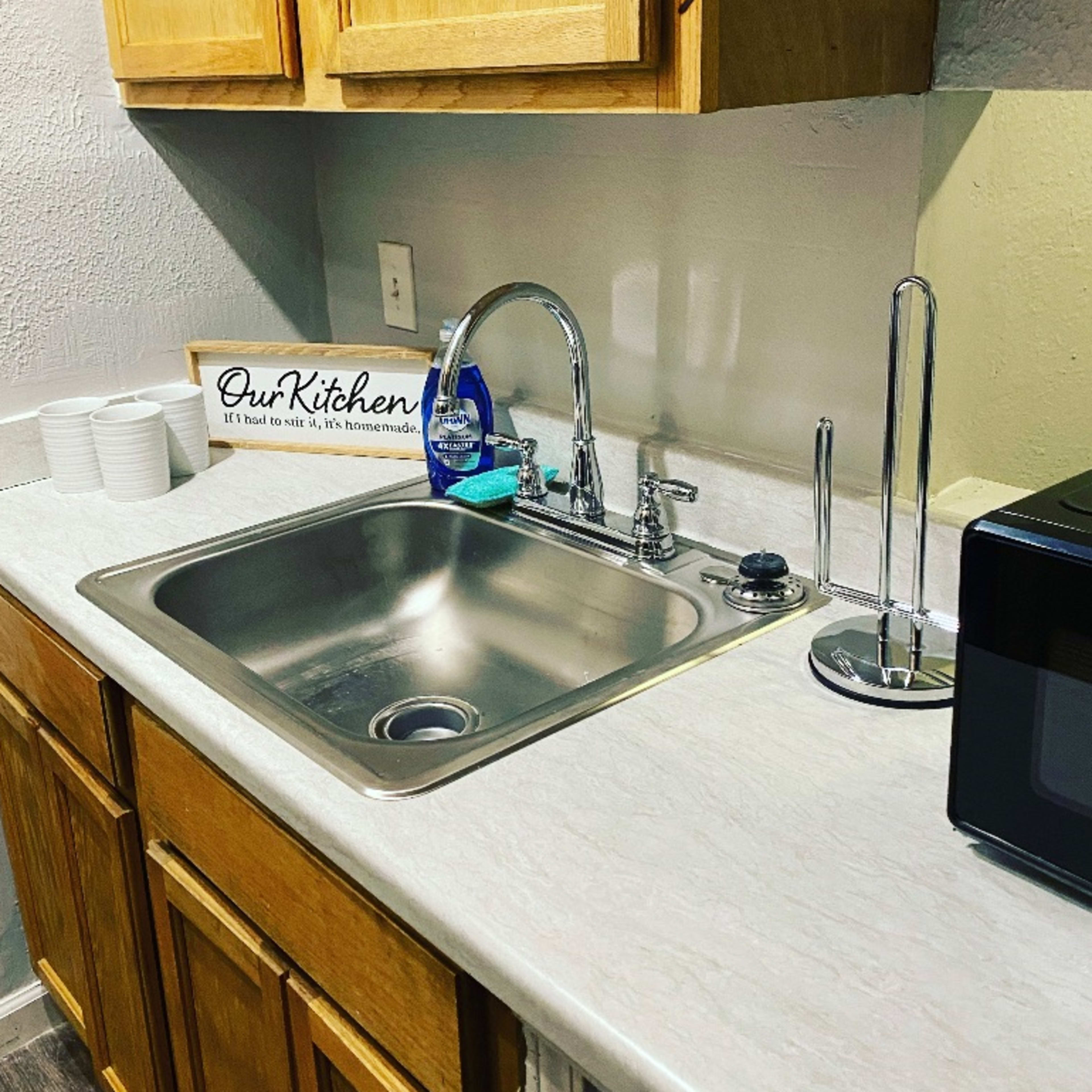 A kitchen sink with a silver faucet, a dish drying rack, a microwave, and wooden cabinets above a light-colored countertop.