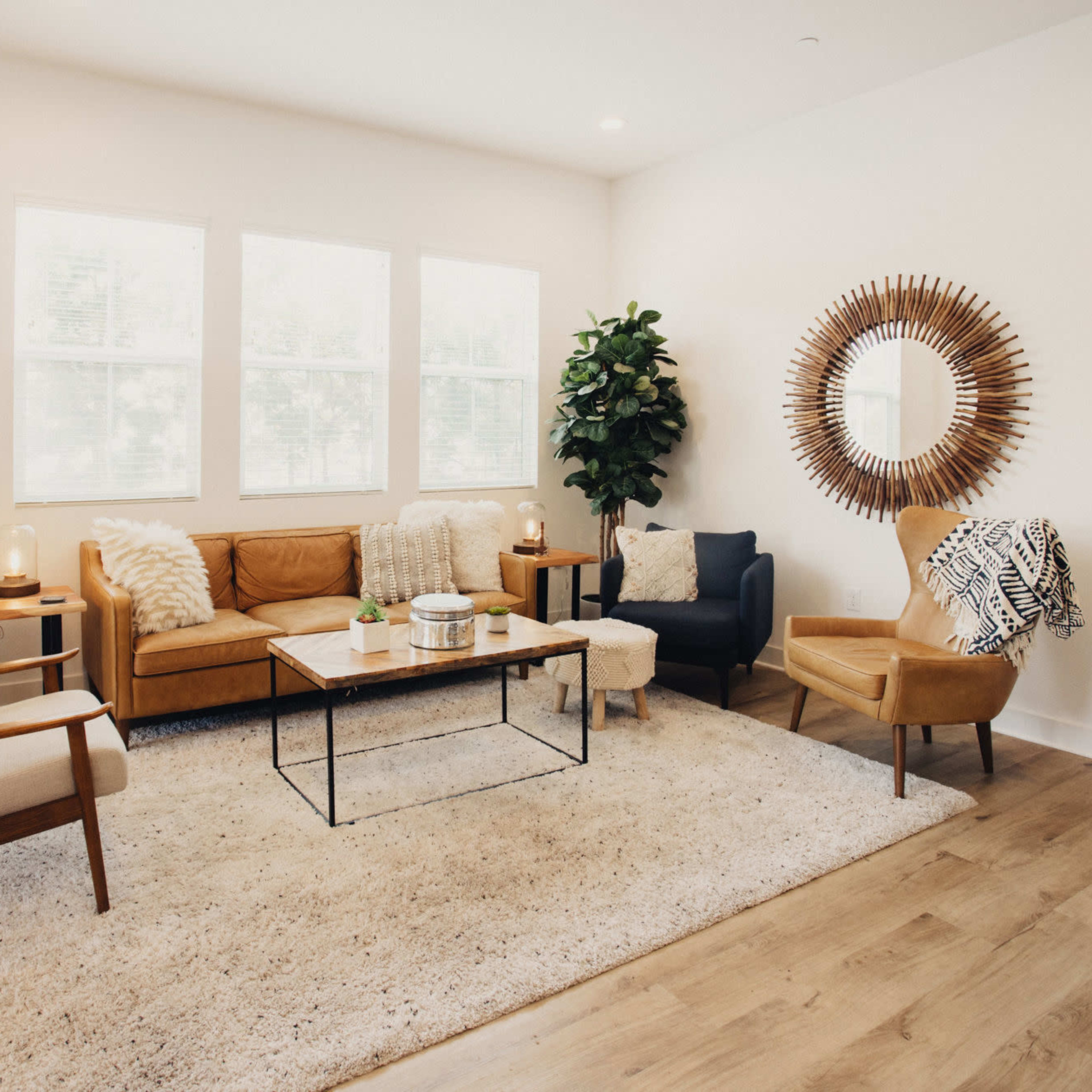 A modern living room furnished with a brown leather sofa, two accent chairs, a coffee table, and a large decorative mirror, all arranged on a light-colored rug.
