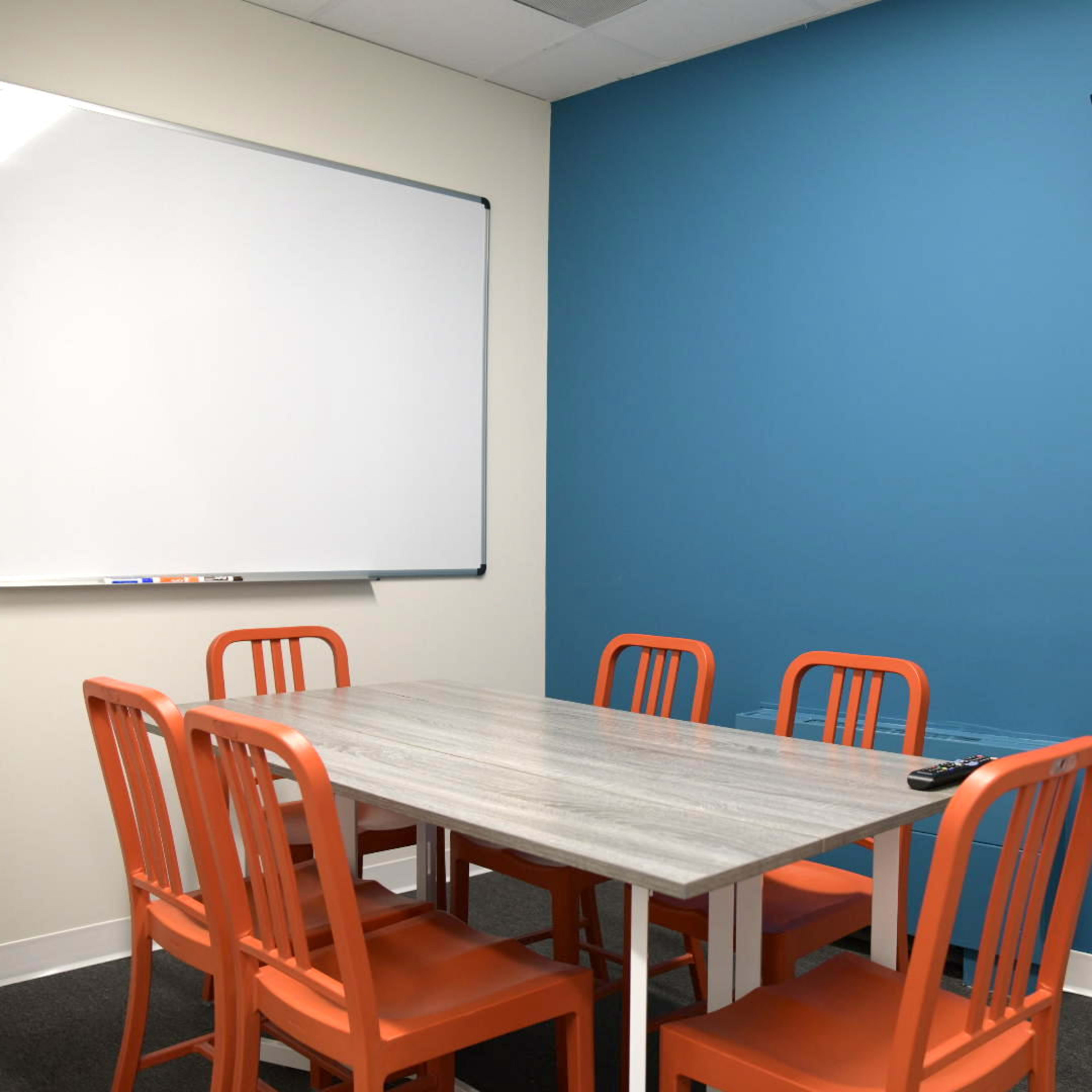 A small meeting room with a gray table, six orange chairs, a whiteboard on one wall, and a mounted television on another wall.
