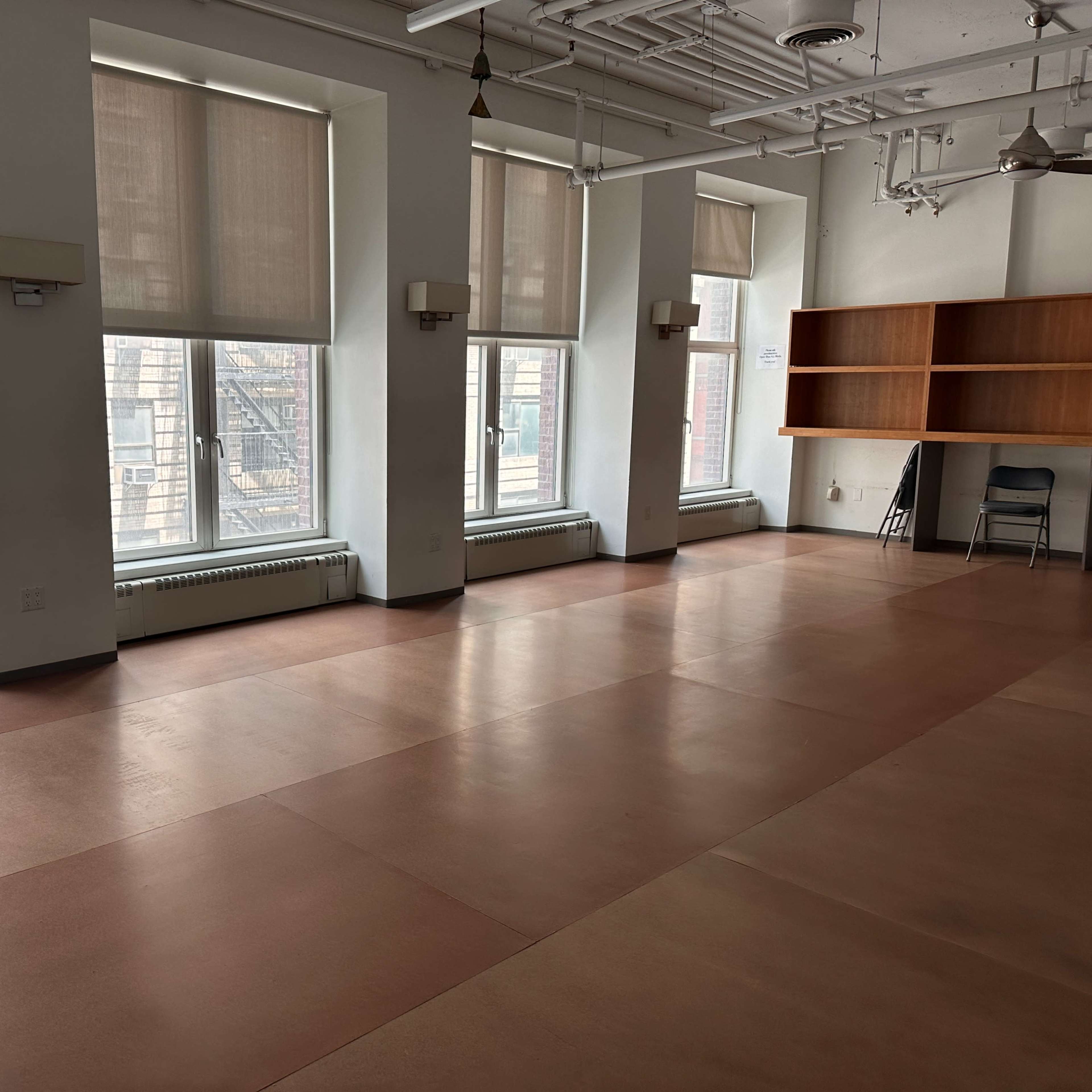 An empty room with large windows, a light-colored floor, and a wooden shelf against one wall.