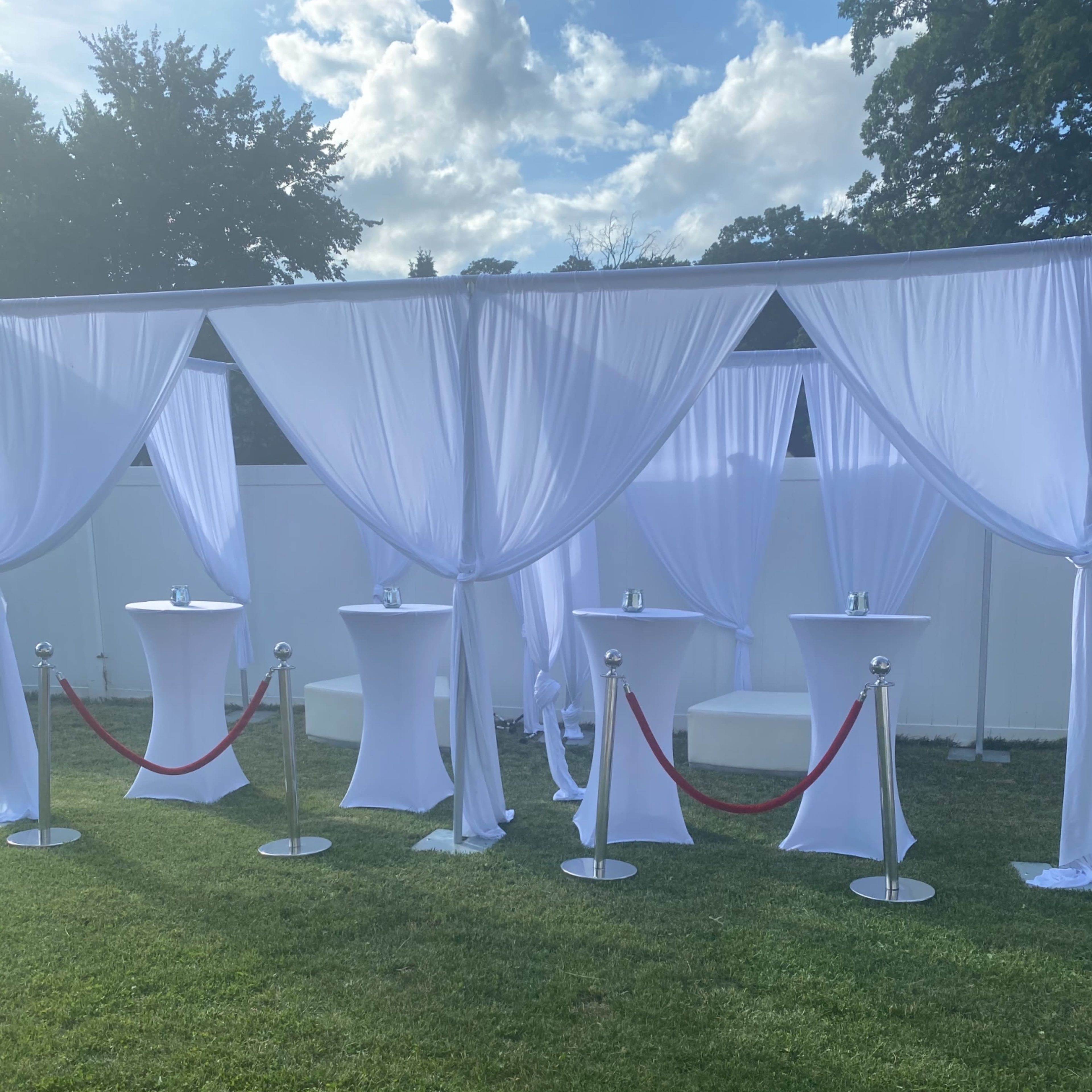 A landscaped area features white draped tents with tall, round tables separated by a red rope barrier.