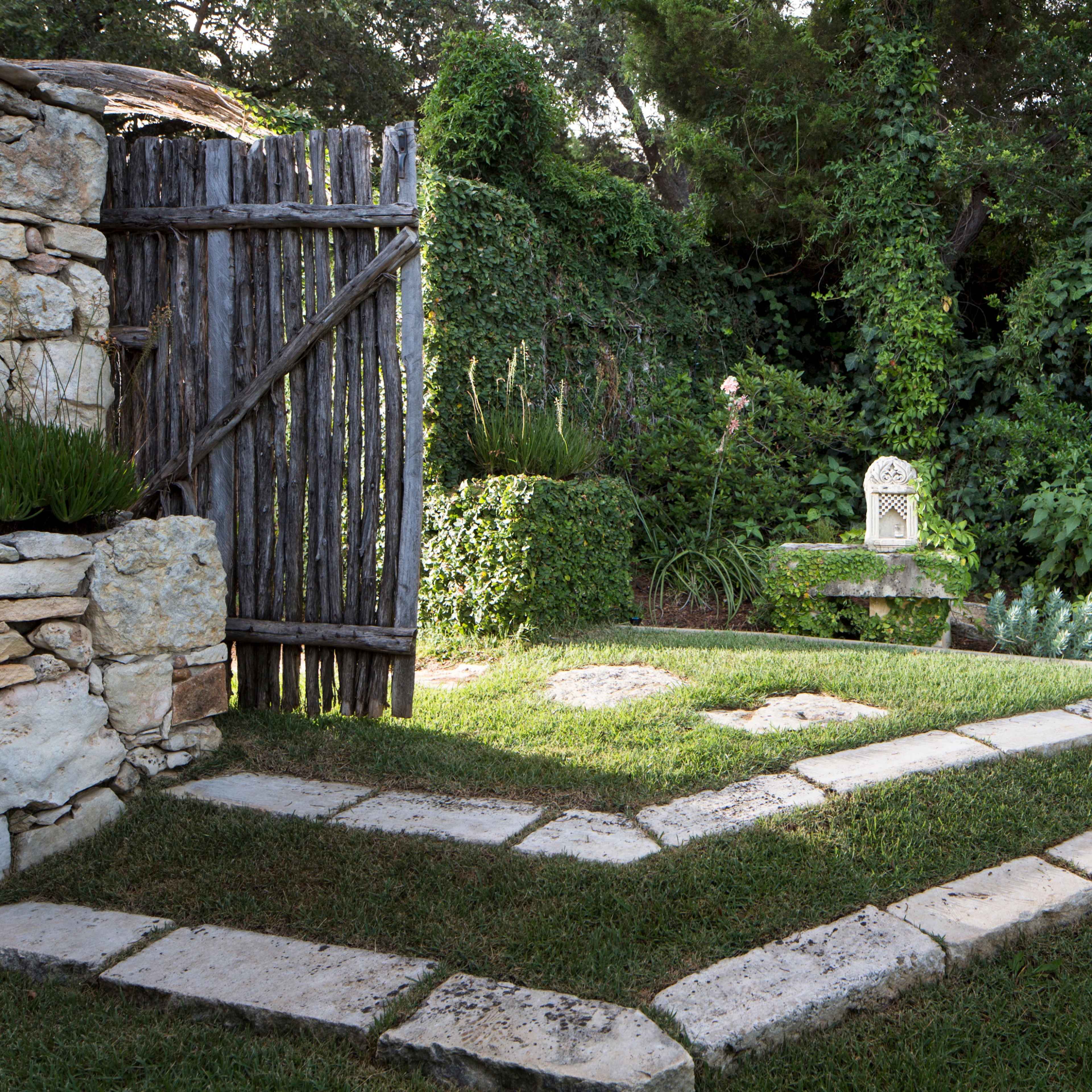 A stone pathway leads through a garden, bordered by lush greenery and a wooden fence.