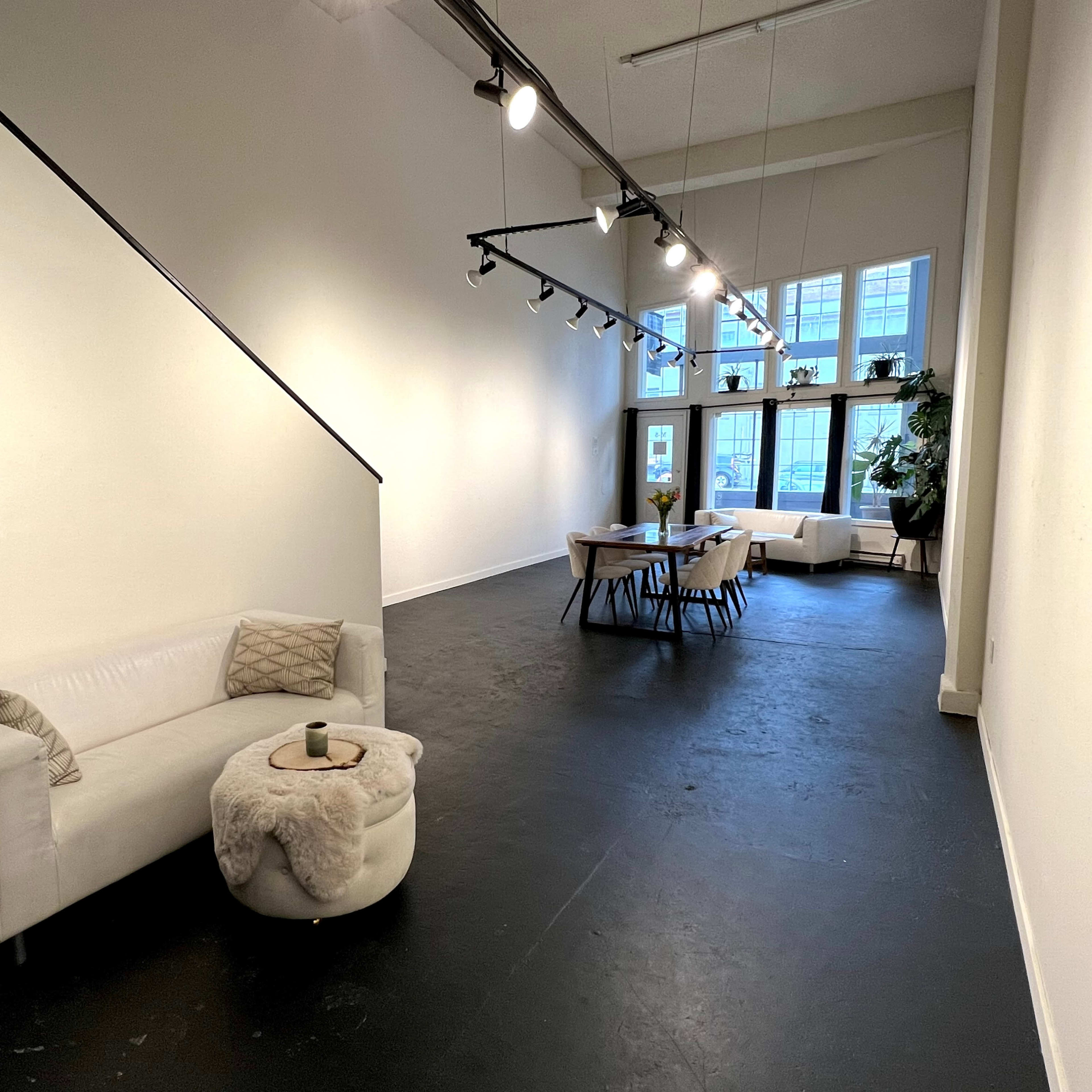 A modern, open interior space featuring a white sofa, a circular ottoman, a large dining table, and large windows allowing natural light.
