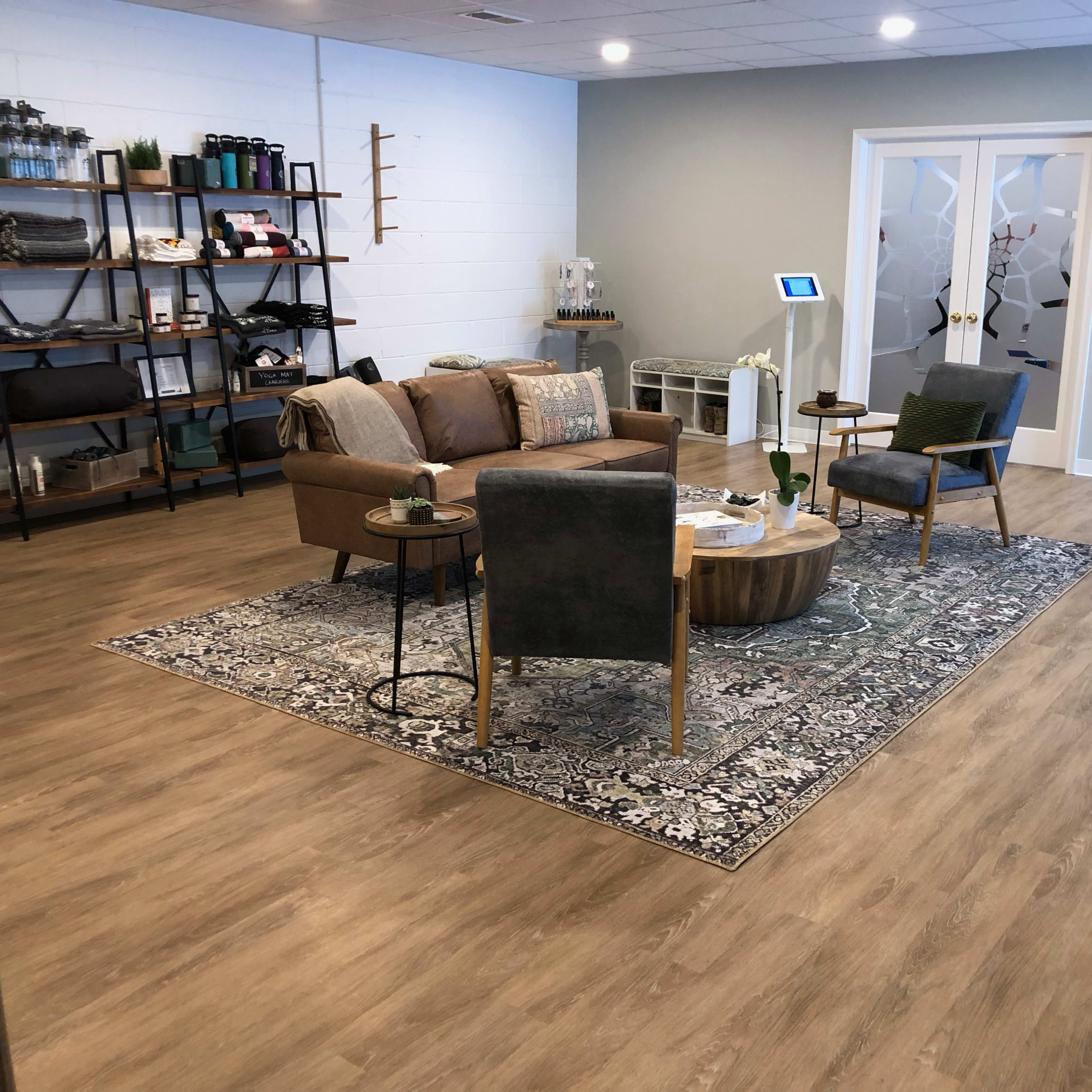 A modern waiting area containing a brown couch, two chairs, a coffee table, and shelves with various items, all arranged on a patterned area rug and wooden floor.