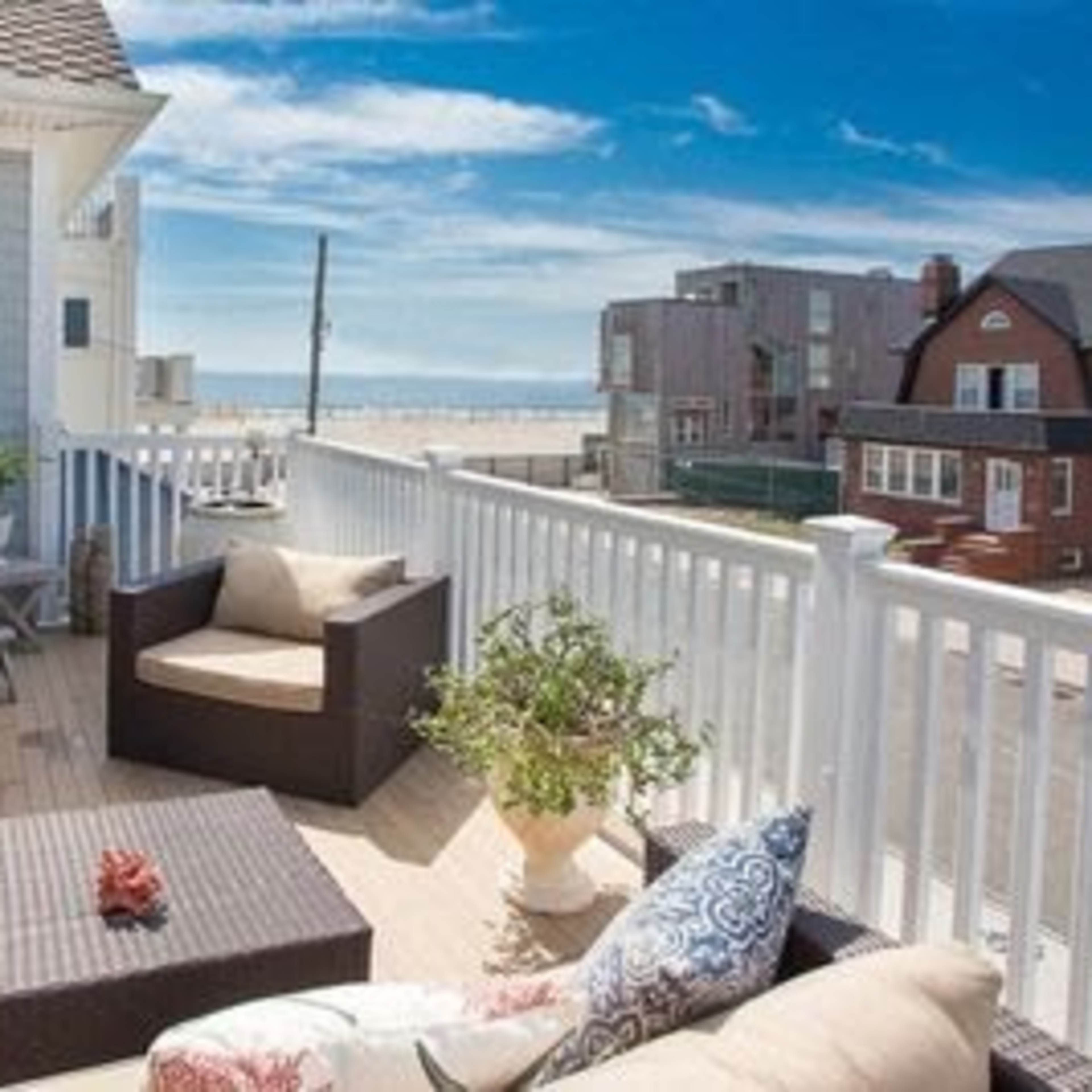 A spacious deck features comfortable seating and plants, overlooking a sandy beach and nearby buildings under a clear sky.