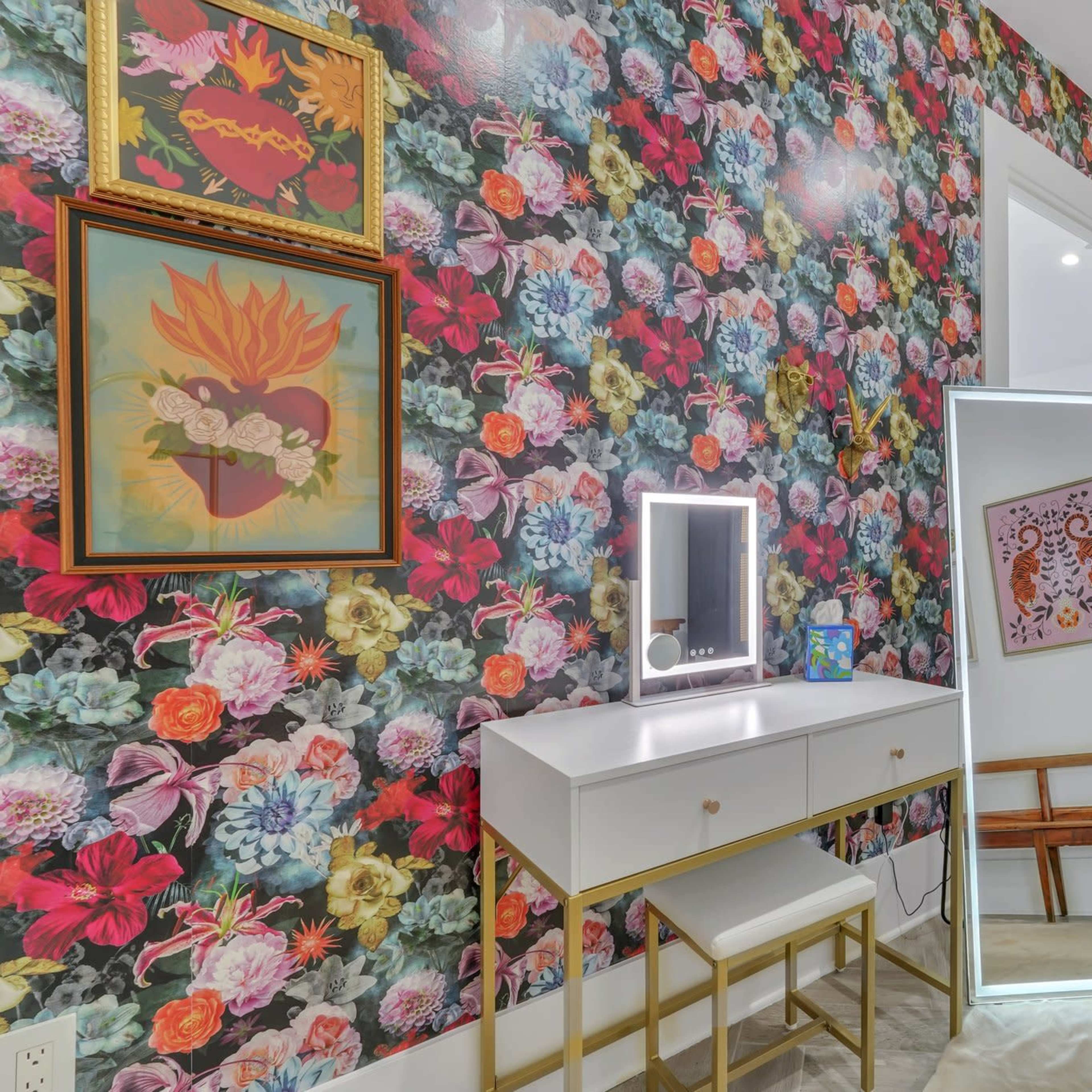 A brightly decorated room featuring a floral wallpaper, a white vanity with a mirror, and a kitchen area in the background.