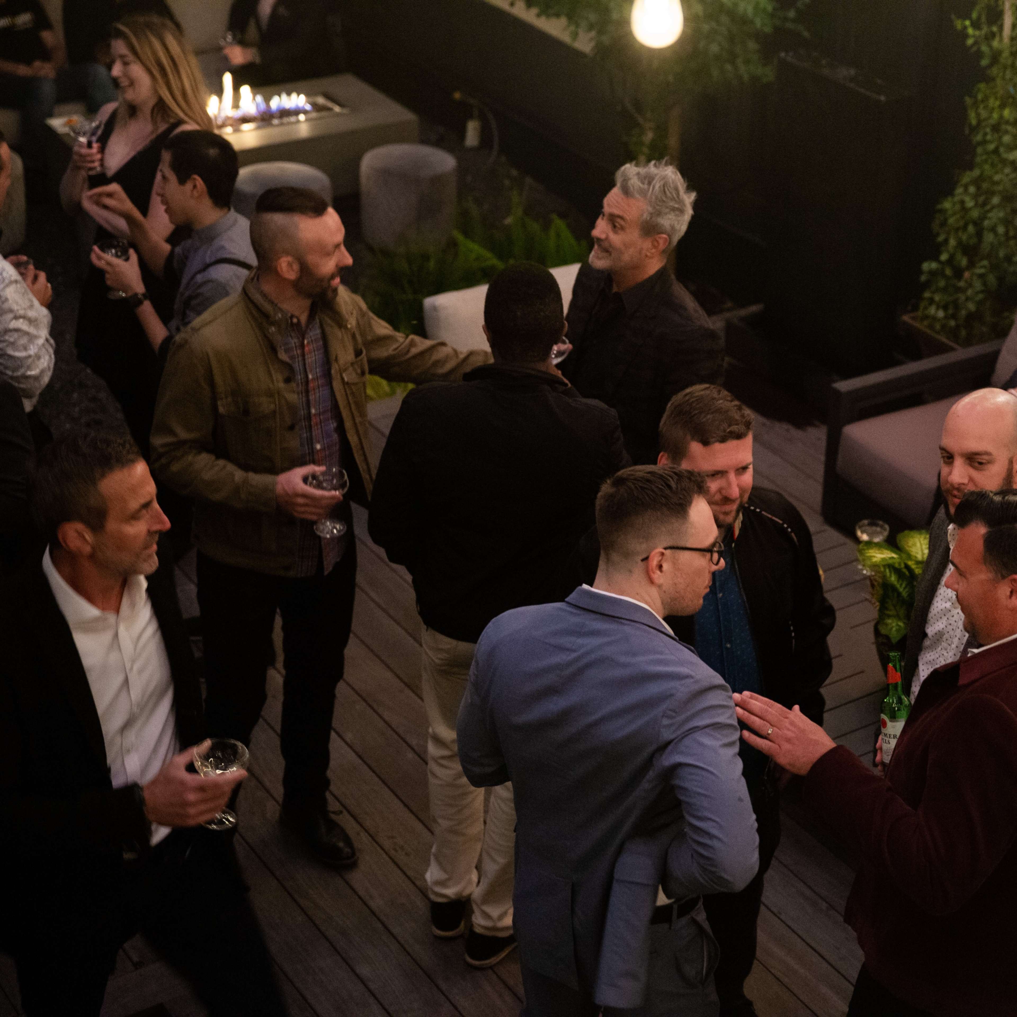 A group of people mingles on an outdoor patio, surrounded by dim lighting and greenery, while others are seated nearby.