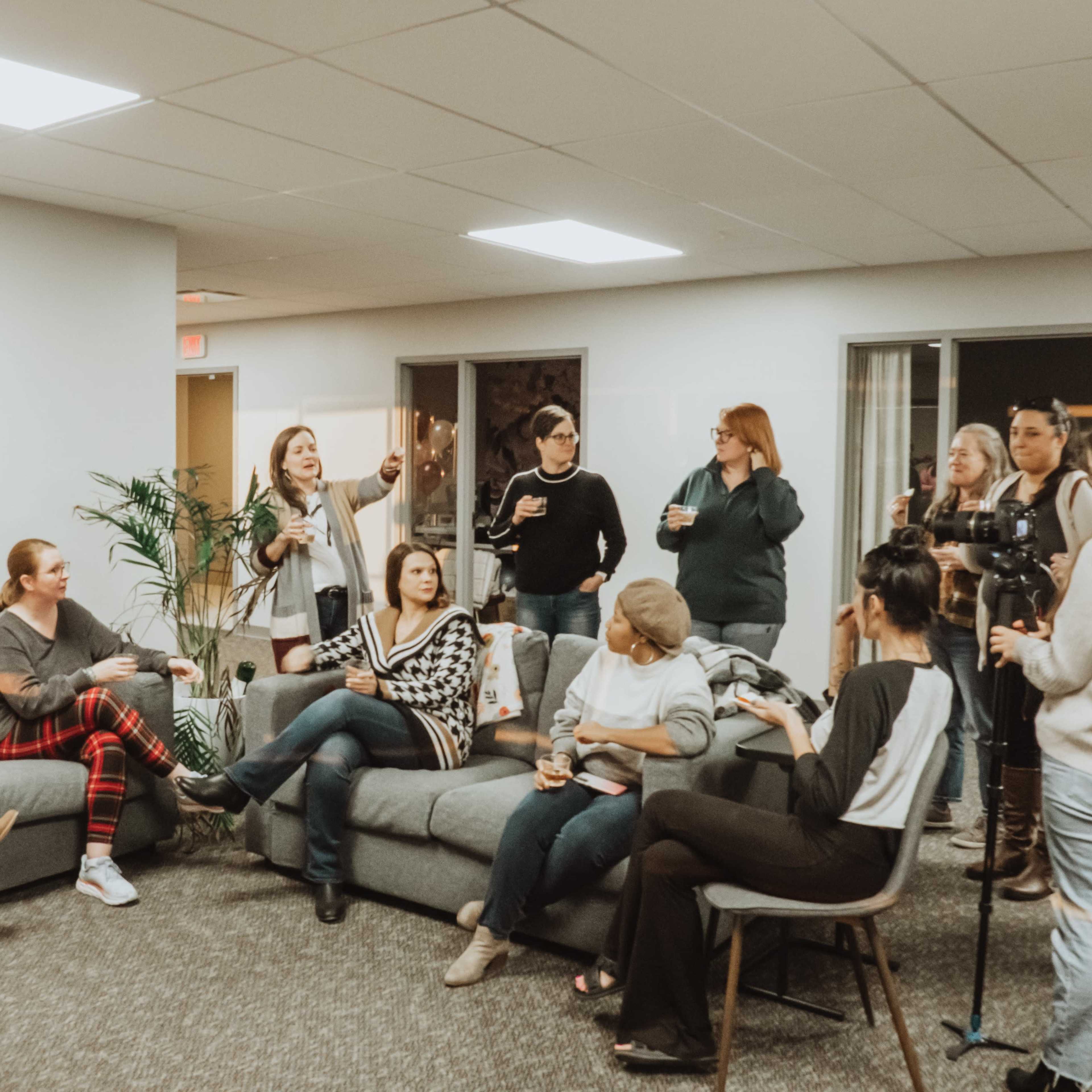 A group of women socializes in a modern office lounge, some standing and others seated on a gray couch.