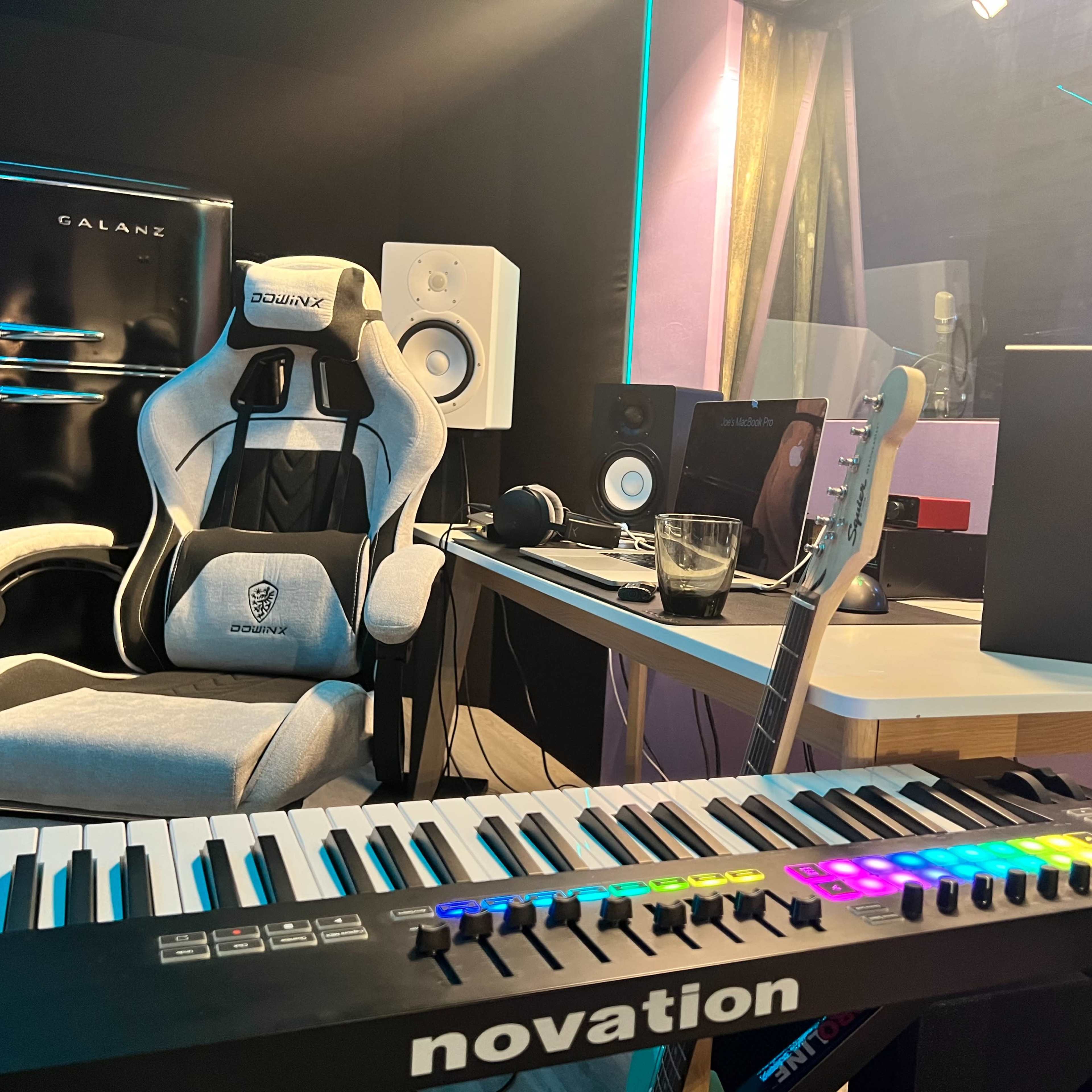 A music production setup featuring a keyboard, a guitar, a desk with speakers, and a gaming chair in a room with purple walls.