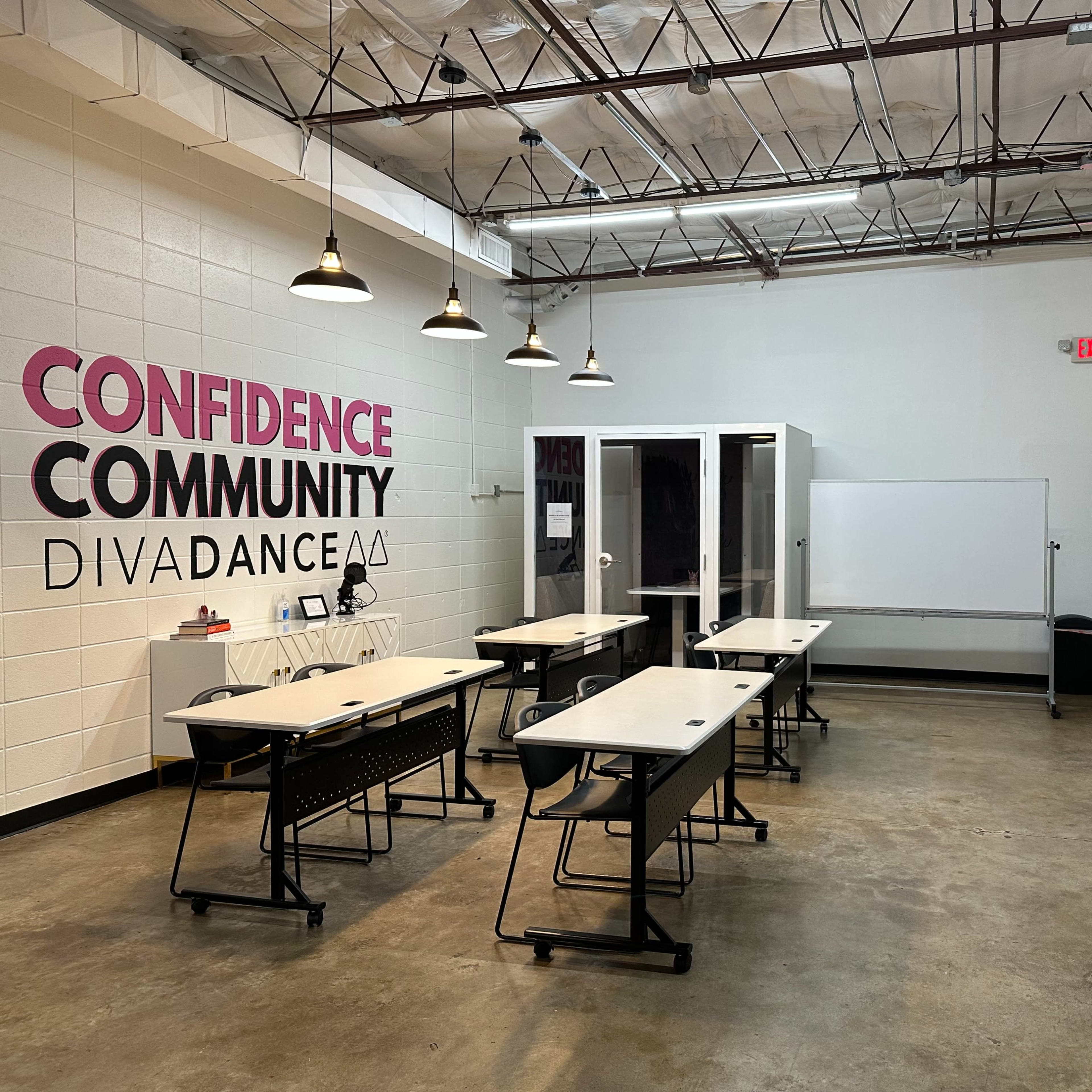 A spacious classroom with several desks arranged in rows, a whiteboard, and a large TV on the wall, with the words "Confidence, Community, DivaDance" displayed prominently.