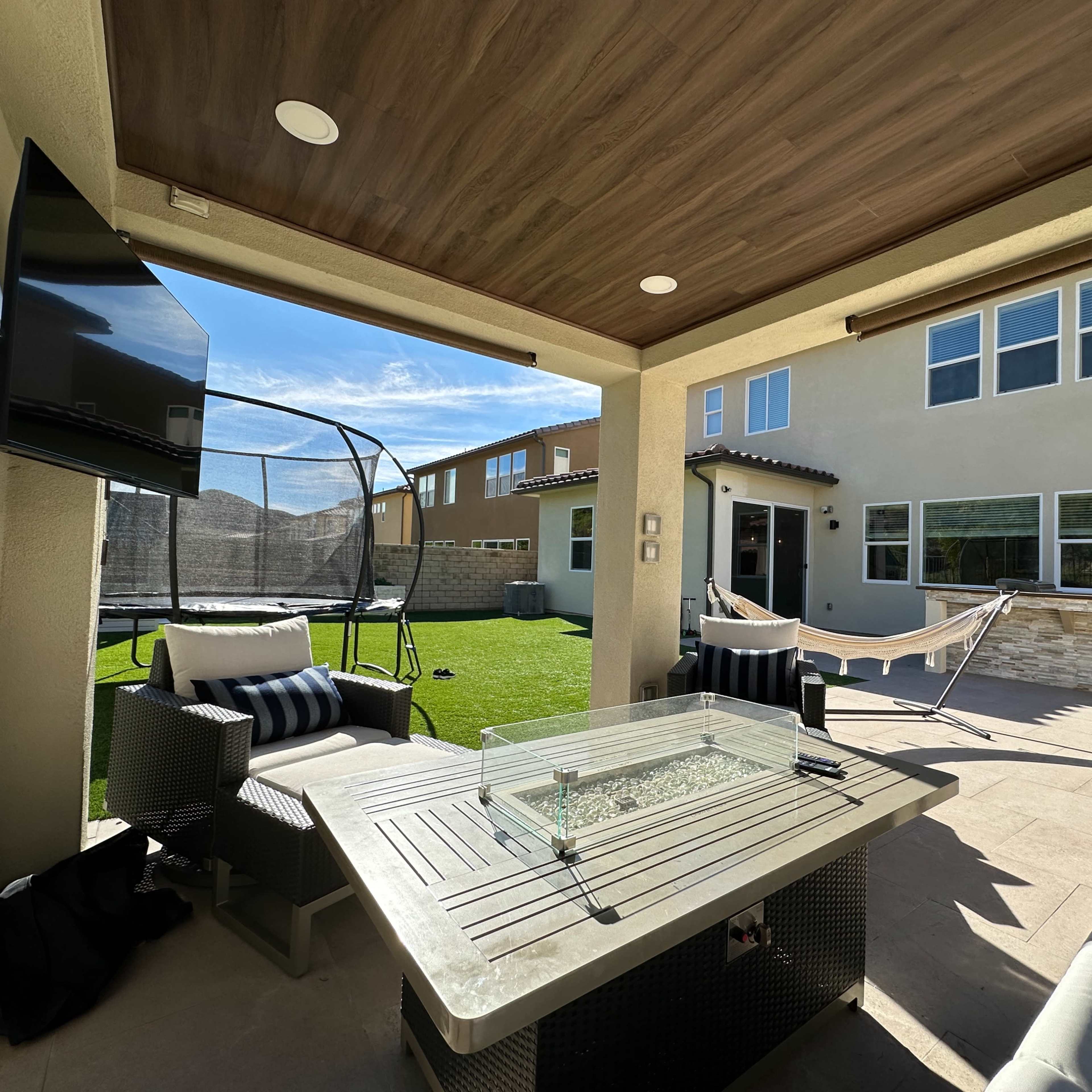 A covered patio area with a fire table, comfortable seating, a television mounted on the wall, and a view of a backyard with a trampoline and hammock.