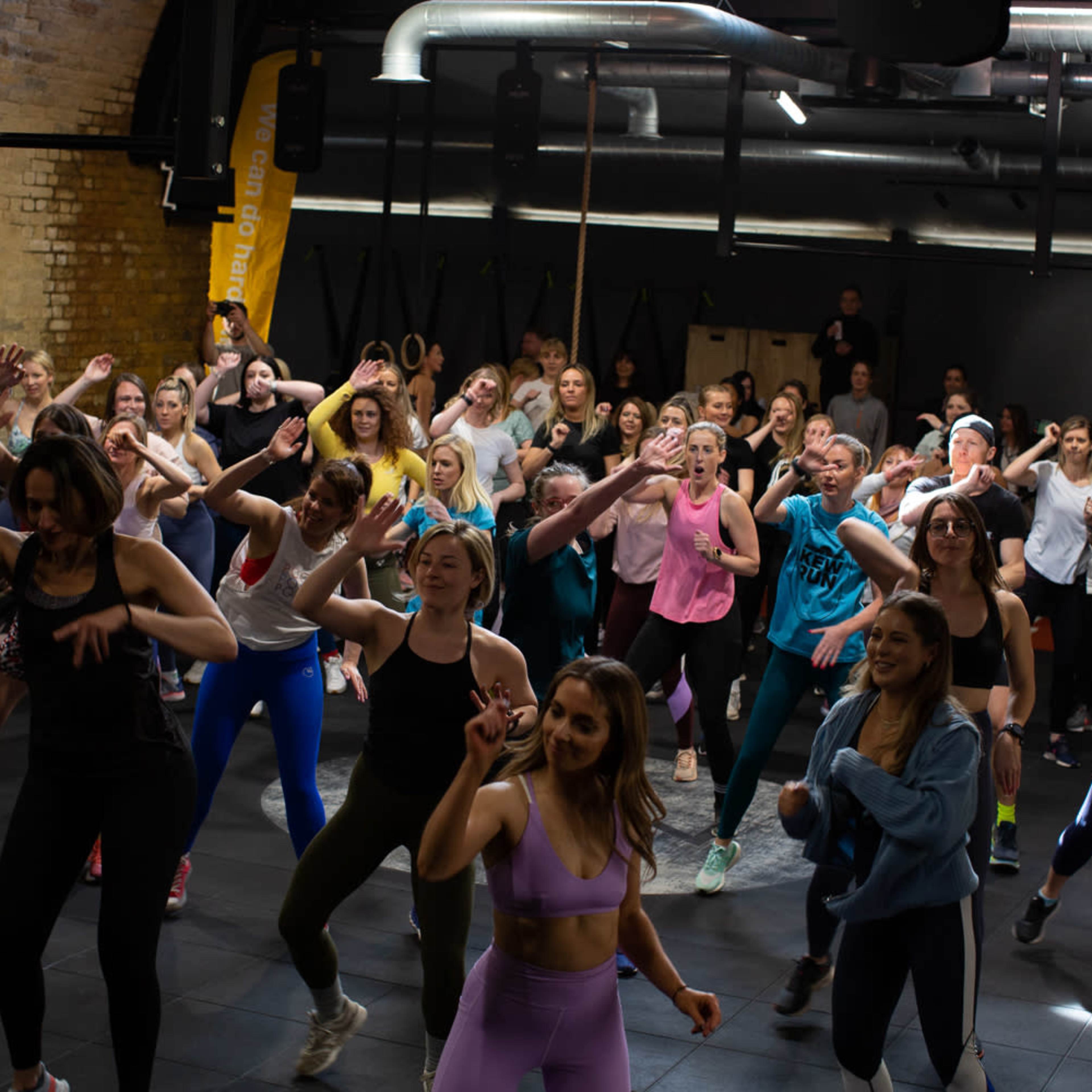 A large group of people is participating in a lively fitness class in a gym, with various individuals engaged in energetic movements.