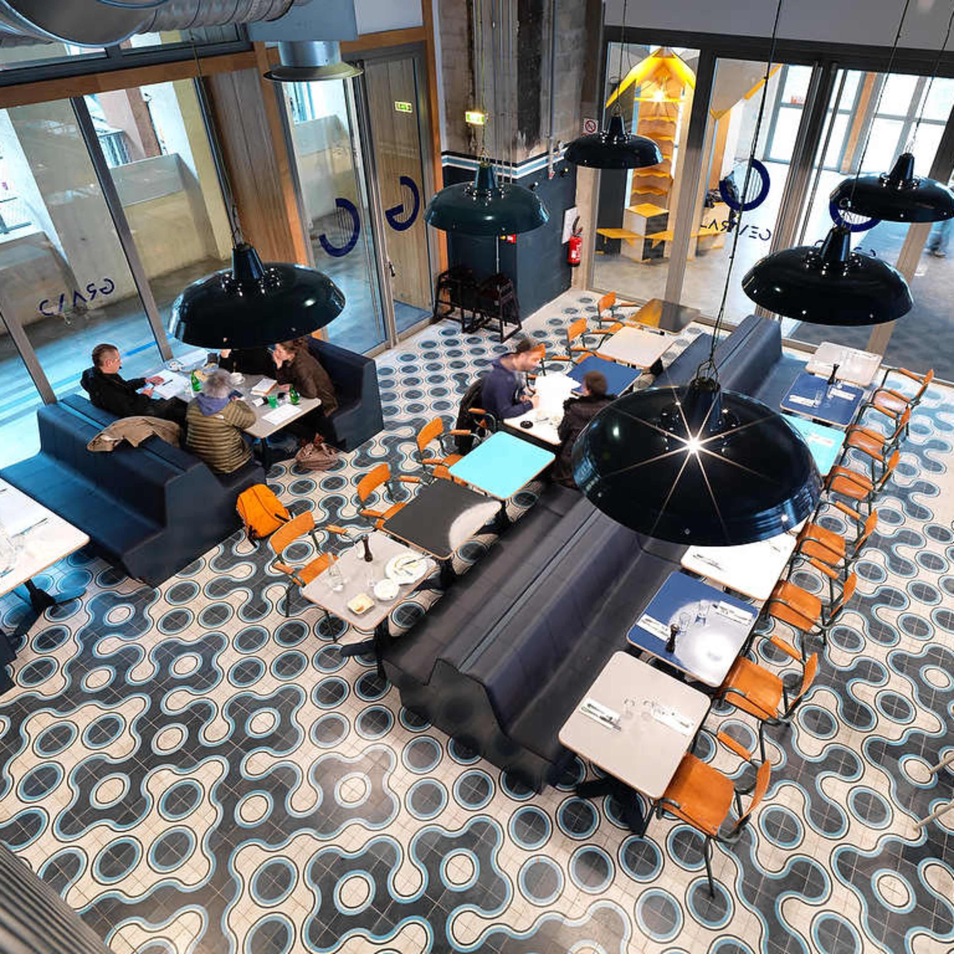 A modern restaurant interior with a patterned tiled floor, large black pendant lights, and a mix of tables and booths filled with diners.