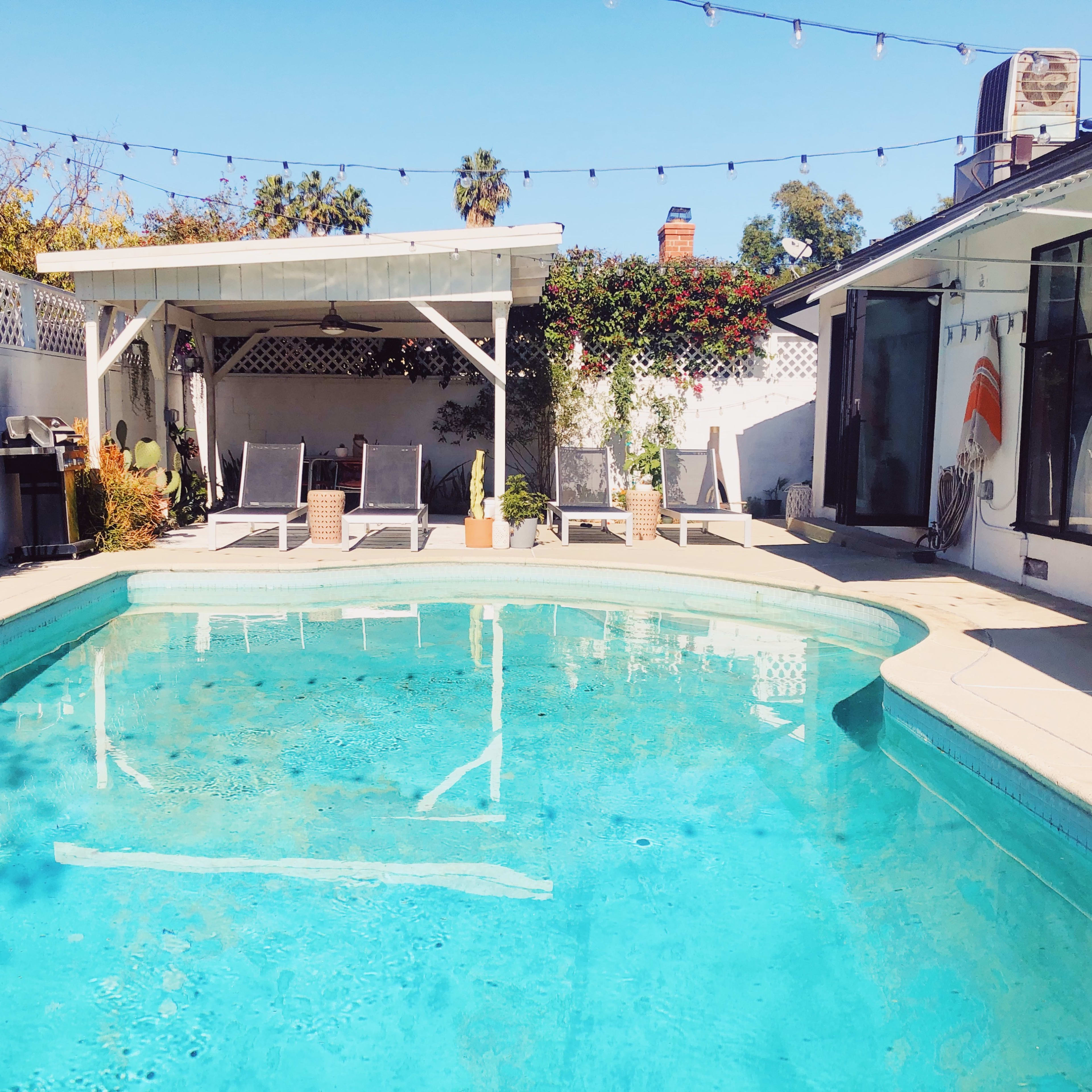 A swimming pool surrounded by lounge chairs, greenery, and a shaded patio area.