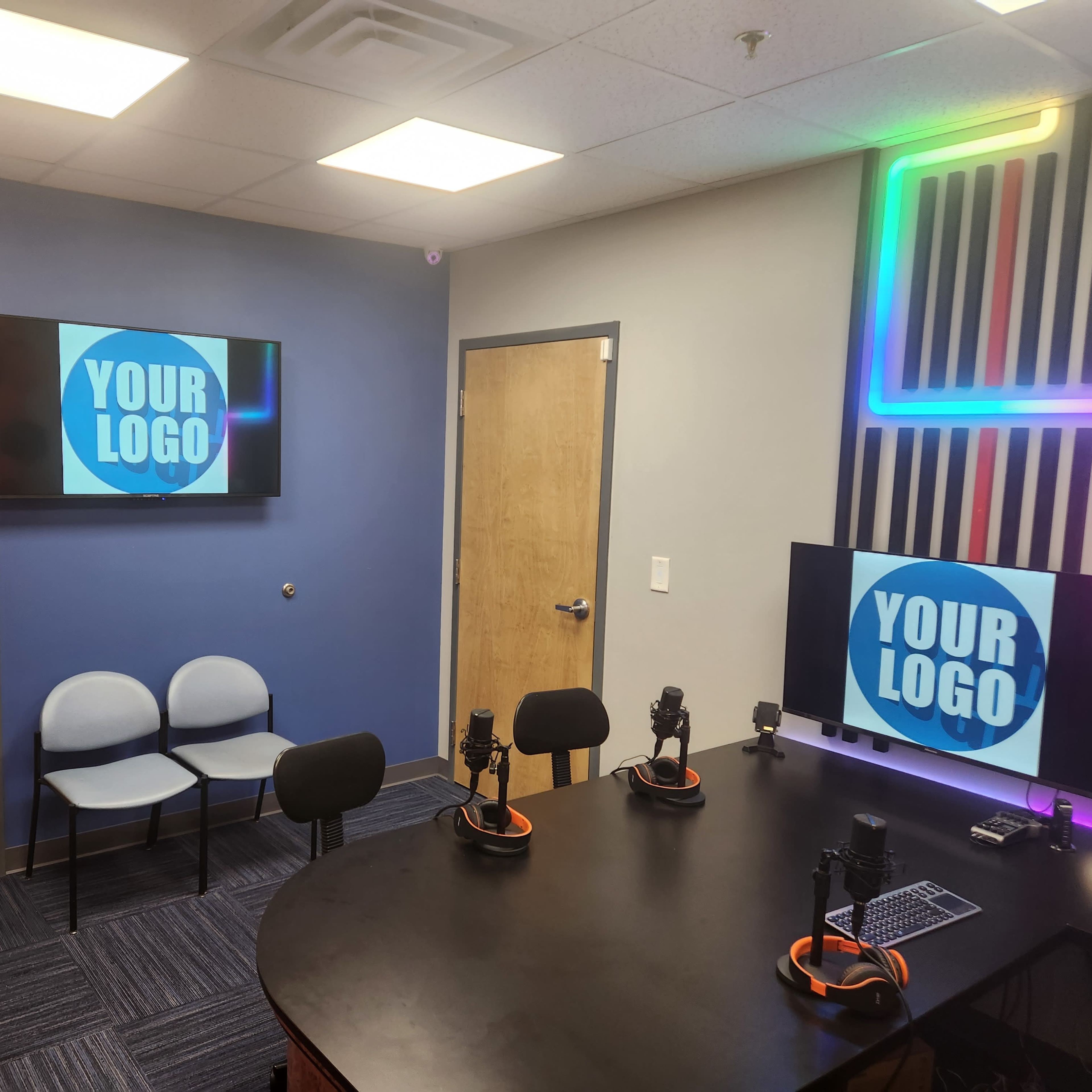 A modern recording studio with two televisions displaying "YOUR LOGO," a round table with microphones, and several chairs arranged nearby.